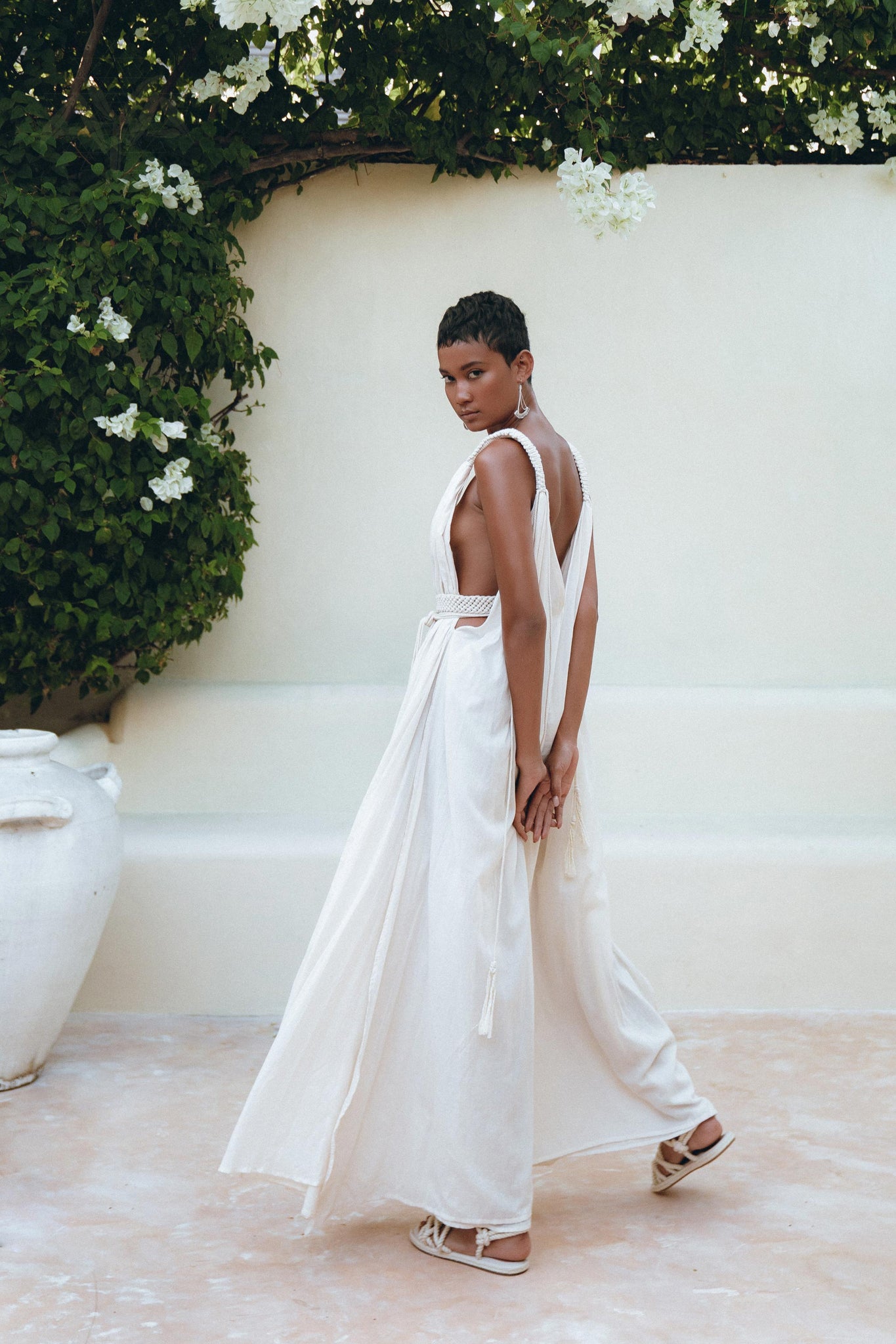Wearing the Boho Beach Macrame Wedding Dress by AYA Sacred Wear, an individual stands gracefully before a cream-colored wall adorned with vibrant green foliage. With a calm expression, they glance over their shoulder. Nearby, a large white ceramic pot adds an artistic touch to the scene.
