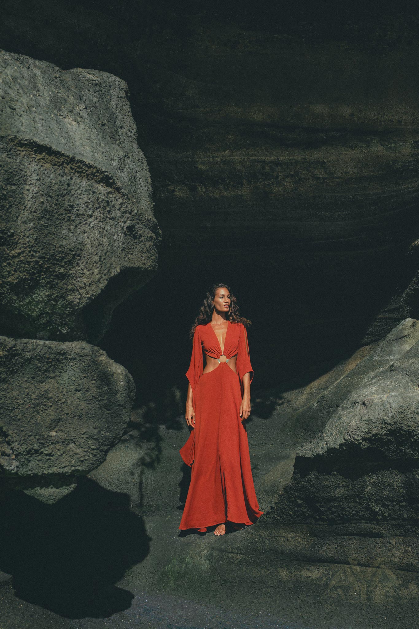 A woman, barefoot with long curly hair, stands between large rock formations wearing an AYA Sacred Wear Red Rose Greek Goddess Dress. Dramatic lighting casts shadows that highlight the dress's elegance and the rocky textures around her.