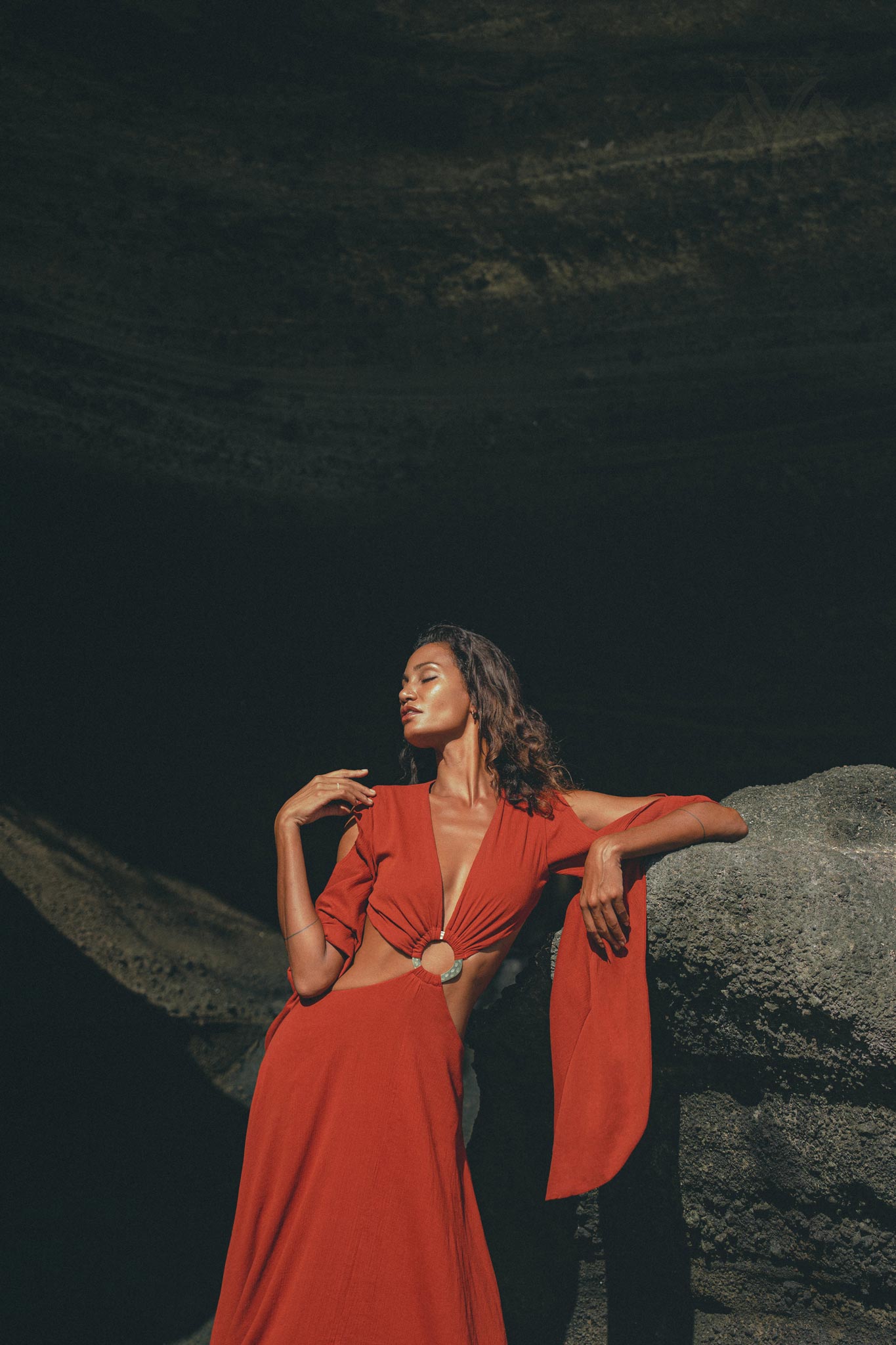 A woman gracefully poses in a Red Rose Greek Goddess Dress by AYA Sacred Wear, featuring bell sleeves and a waist cutout. She stands against rock formations, with her hand gently on a rock and eyes closed, sunlight illuminating her and the artisanal cotton gown.