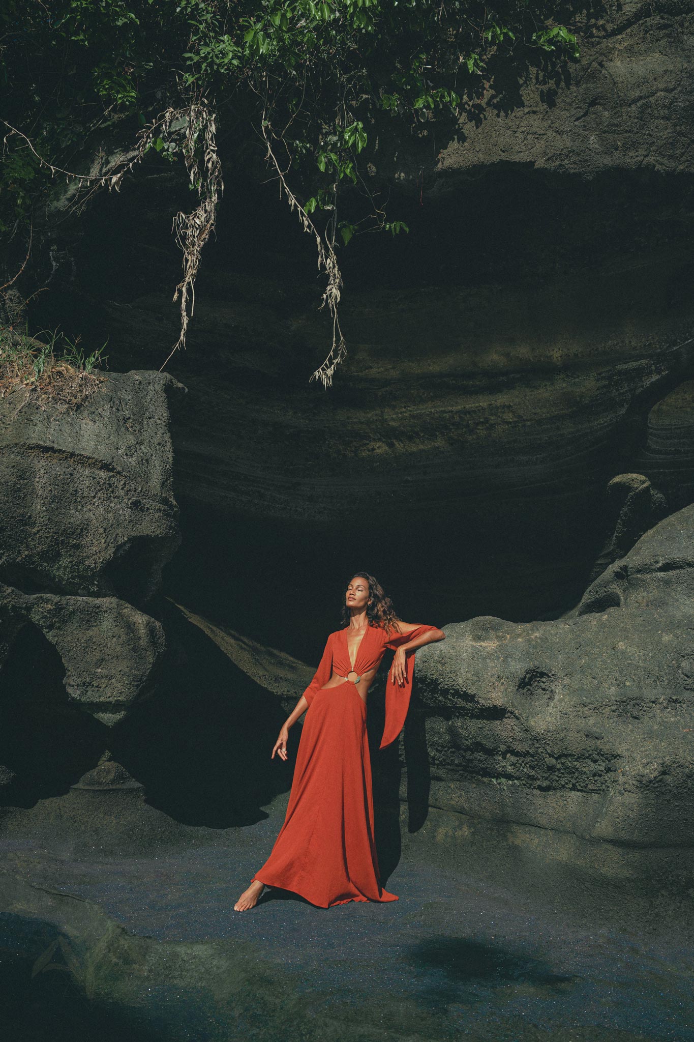 A woman in an AYA Sacred Wear Red Rose Greek Goddess Dress poses gracefully against a rocky backdrop. The lighting accentuates her, casting shadows on the rocks. Lush greenery hangs above, enhancing the enchanting scene.