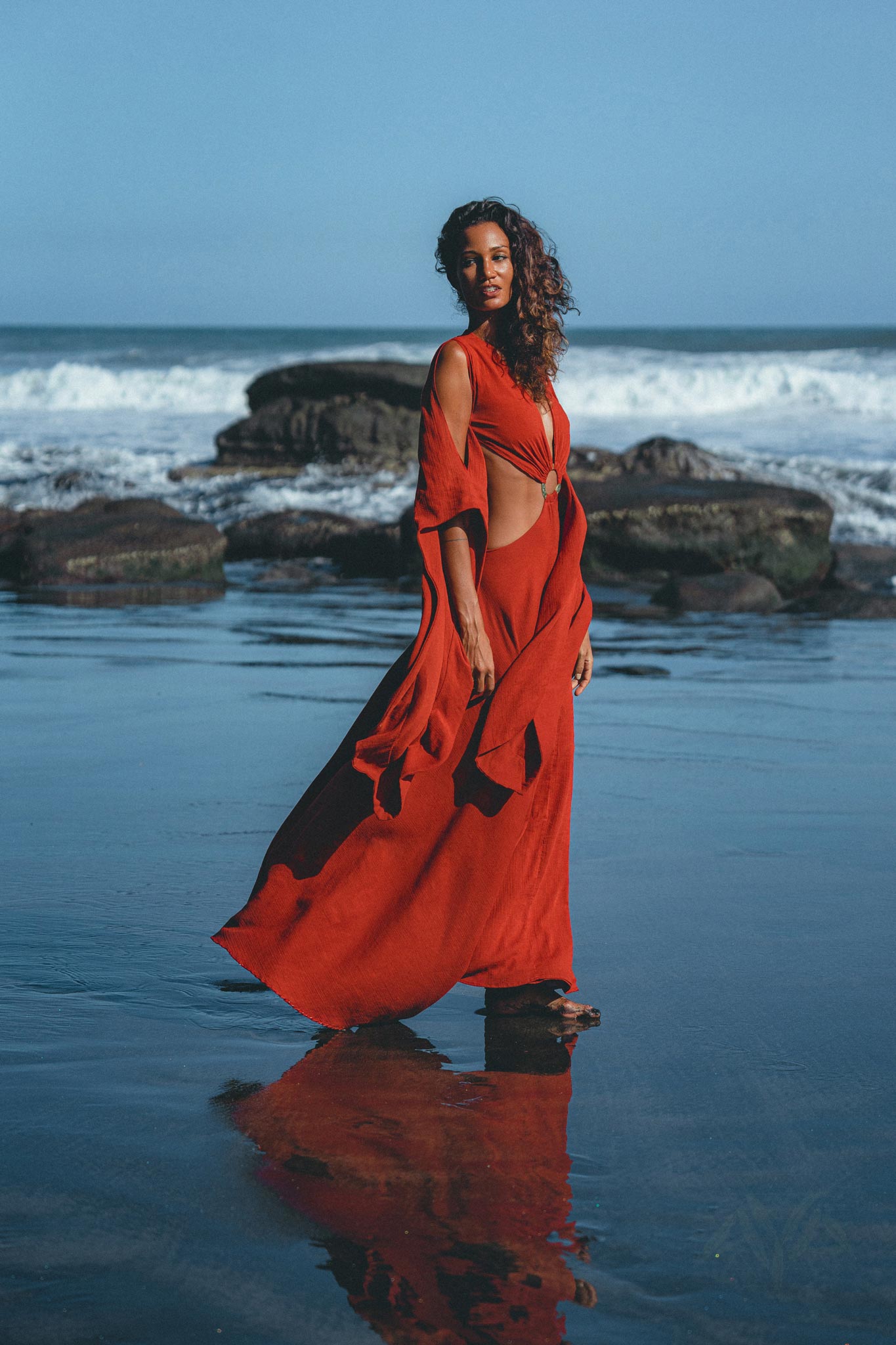 A woman in an AYA Sacred Wear Red Rose Greek Goddess Dress stands confidently on a beach, as waves crash on rocks and her reflection graces the wet sand. Her long, curly hair flows elegantly in the clear sky's backdrop.