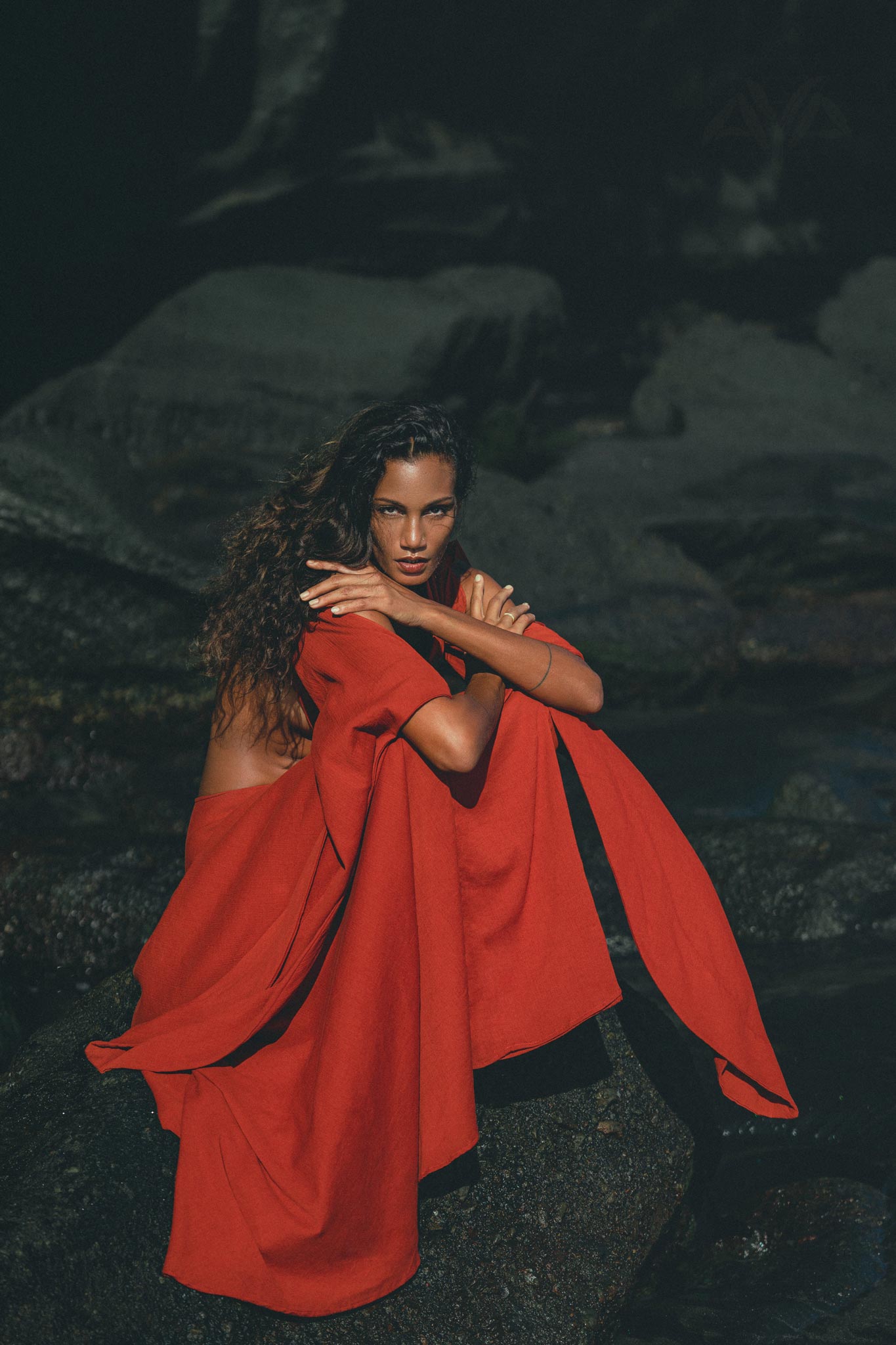 A person with long, wavy hair sits on a rock in the AYA Sacred Wear Red Rose Greek Goddess Dress. Their arms are crossed, gazing at the camera with dark stones in the background, adding a dramatic touch.
