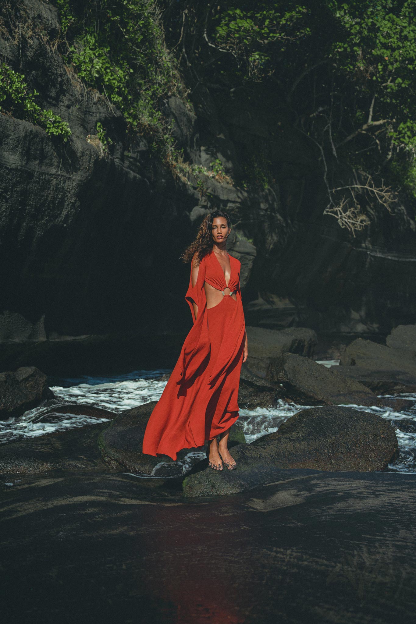 A woman in a flowing Red Rose Greek Goddess Dress by AYA Sacred Wear stands barefoot on rocks by the ocean. Her curly hair is loose in the breeze, with dark cliffs and lush greenery in the background and waves gently crashing around her.