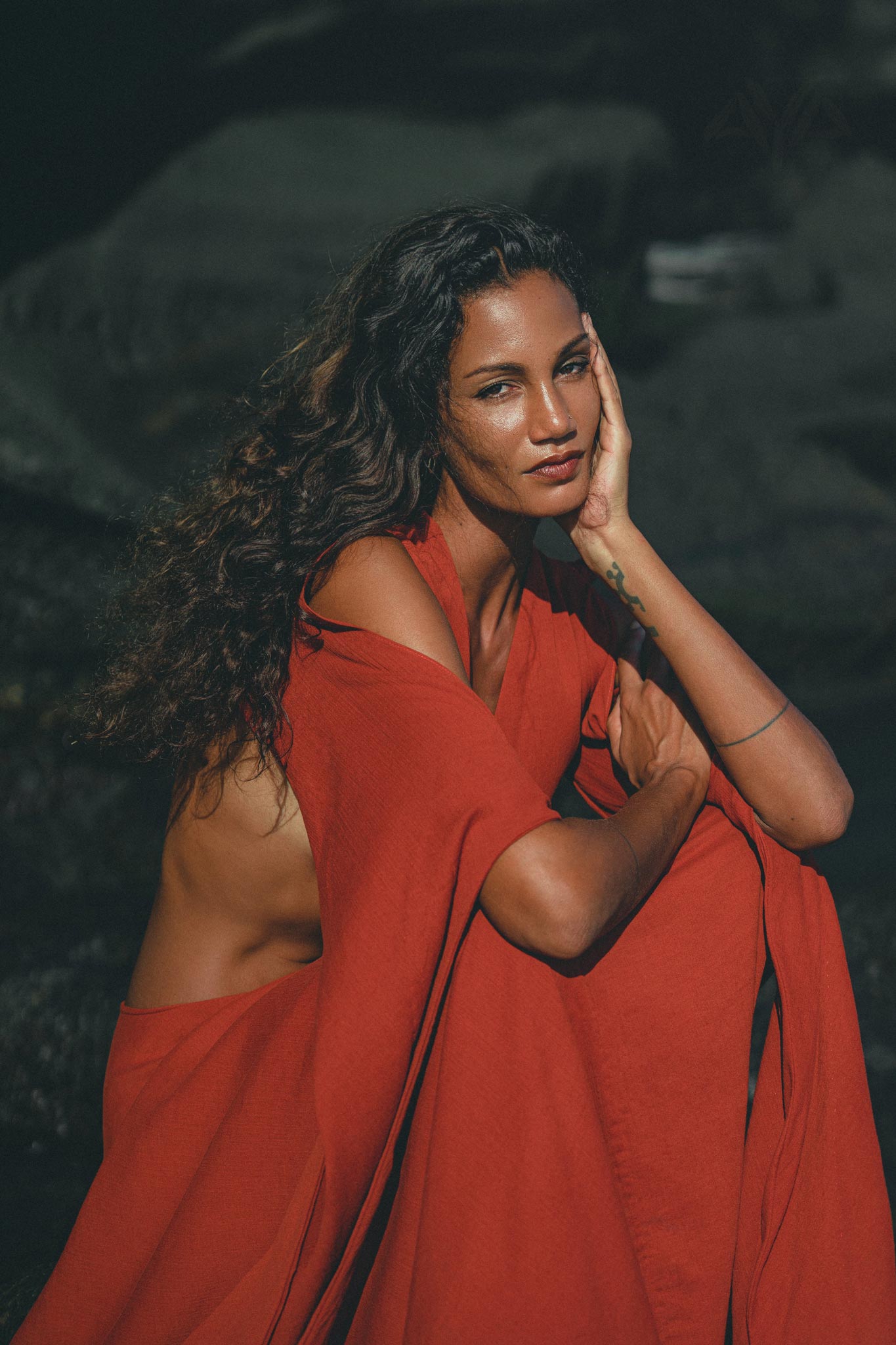 Outdoors, a person dons the Red Rose Greek Goddess Dress by AYA Sacred Wear. Their long, wavy hair and thoughtful expression enhance the bohemian charm of the flowing dress with bell sleeves, set against a dark, natural background.