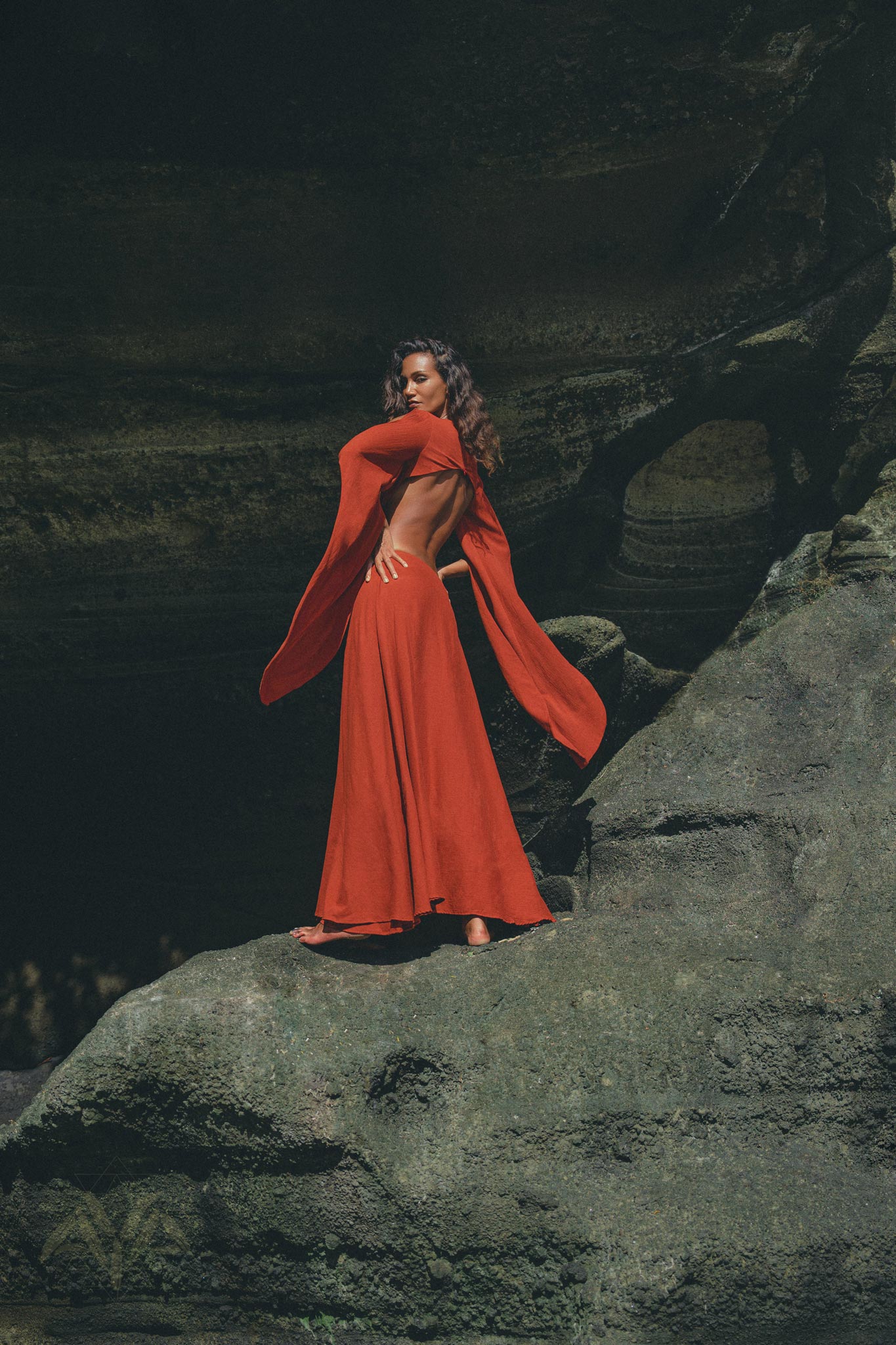 A woman confidently poses in a Red Rose Greek Goddess Dress with bell sleeves by AYA Sacred Wear on a rocky surface. Her artisanal cotton gown trails behind her, contrasting elegantly against the dark, textured rock, creating a dramatic scene.