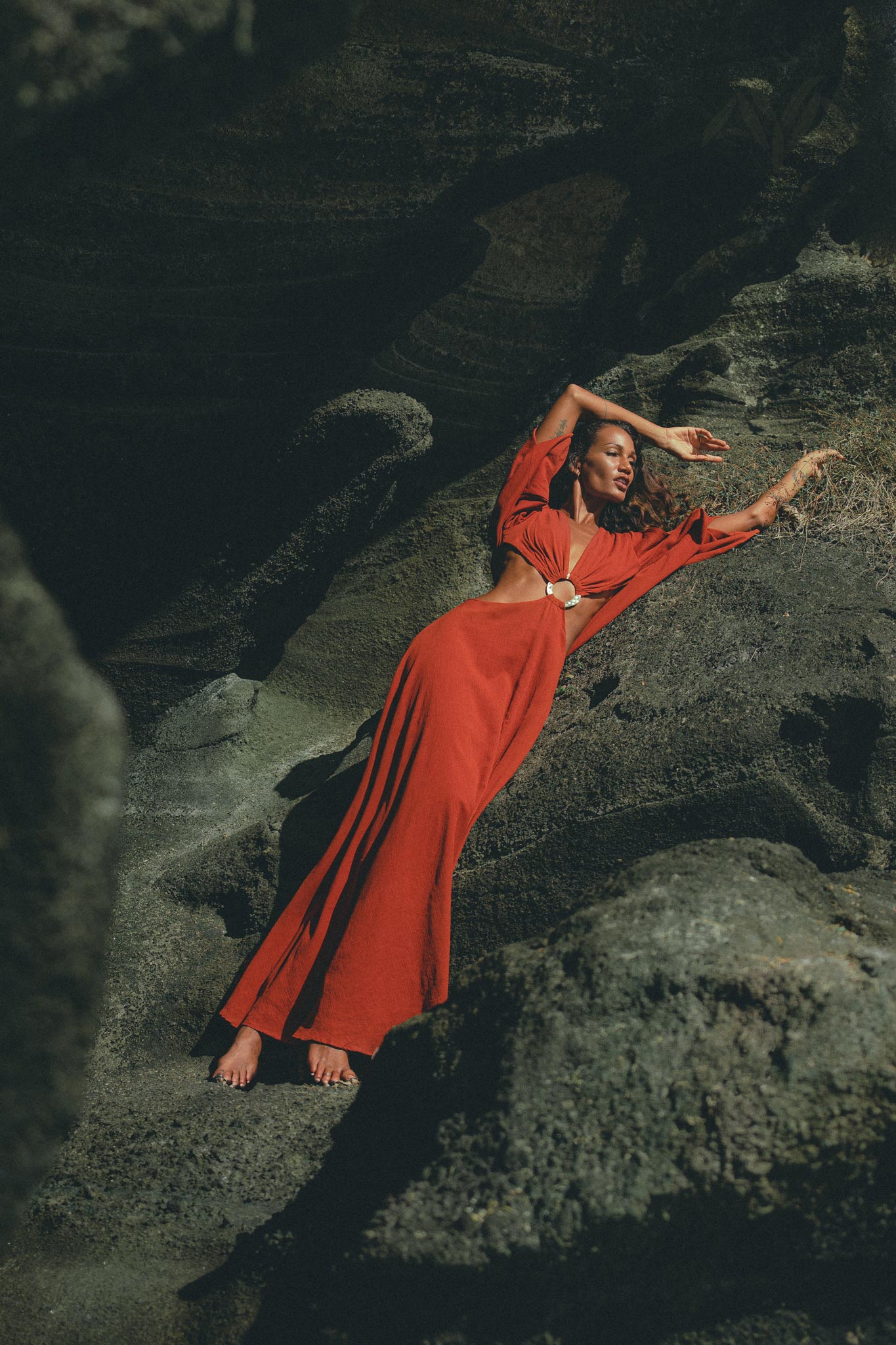 Wearing the Red Rose Greek Goddess Dress by AYA Sacred Wear, featuring bell sleeves and waist cut-outs, a person reclines on rocky terrain. The artisanal cotton fabric's rust color creates a dramatic contrast with the dark setting.