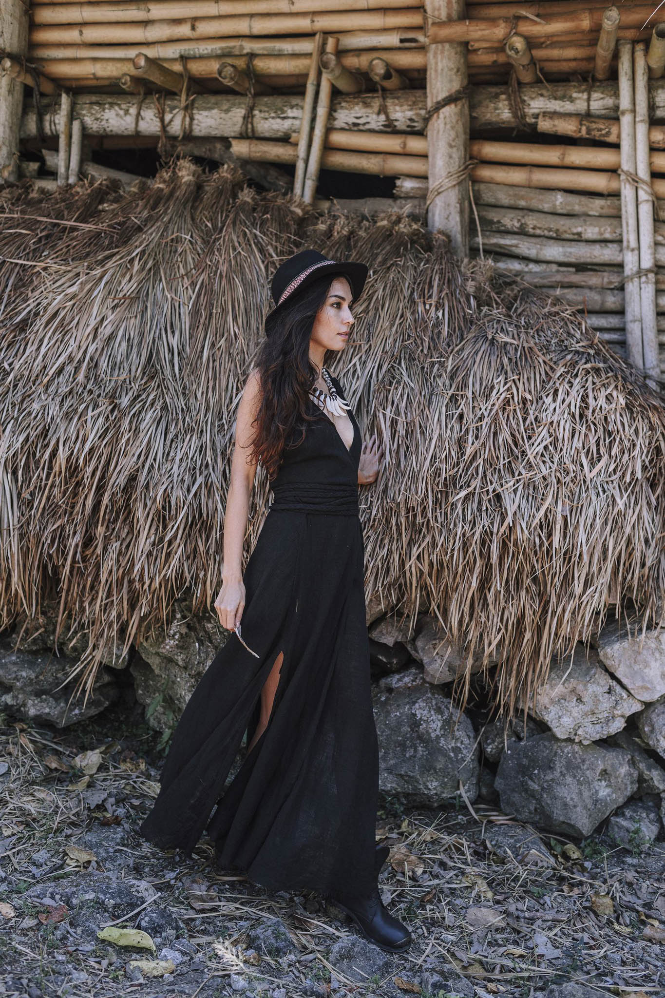 A woman in the Black Cotton Bohemian Dress by AYA Sacred Wear and a hat stands outdoors beside a rustic wooden and dried grass structure, with rocks and leaves scattered on the ground.