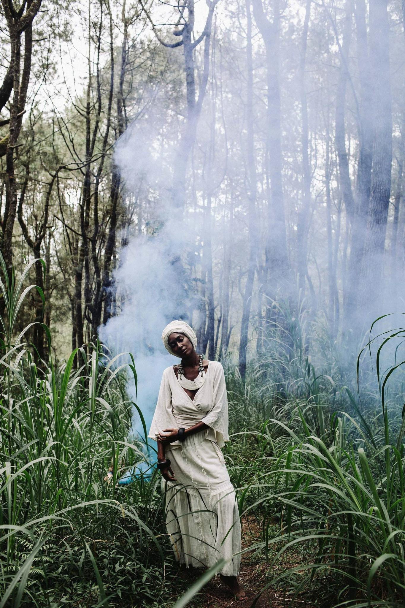 A person dressed in an Off-White Long Boho Skirt with Pockets from AYA Sacred Wear and a headscarf stands amidst tall green grass and trees. A light haze of smoke or mist creates a mystical ambiance in the forest setting. The person appears to be in a serene and contemplative state.