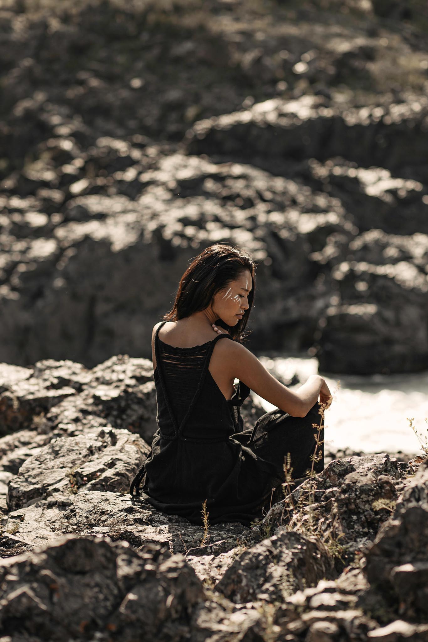 A woman in an AYA Sacred Wear Black Cotton Bohemian Dress sits on rocky terrain, facing away but glancing over her shoulder as sunlight illuminates her face and the textured rocks around her.