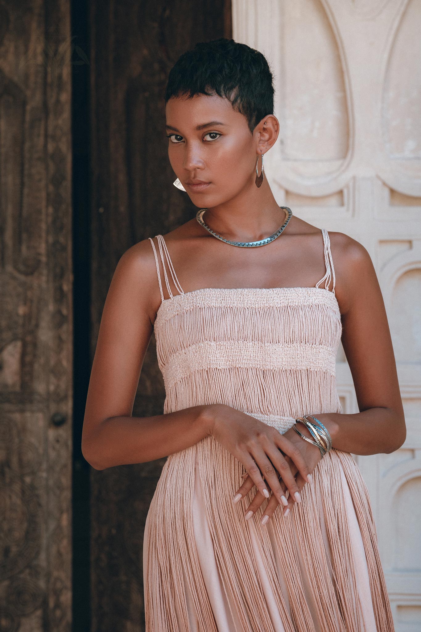 A woman with short hair wears the Dusty Pink Tribal Goddess Slip Over net overlay dress., an ethnic handwoven macrame layer-up sheer net by AYA Sacred Wear. She stands confidently before ornately carved wooden doors, facing the camera.