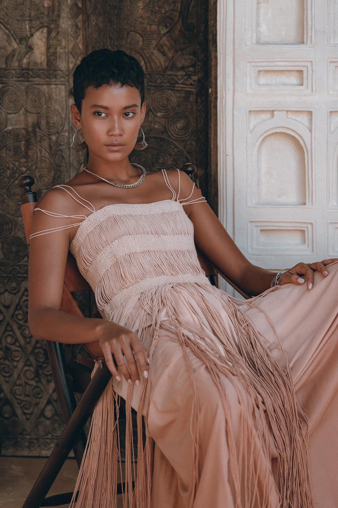 A woman with short hair sits on a chair, wearing the AYA Sacred Wear Dusty Pink Tribal Goddess Slip Over net overlay dress. —an ethnic handwoven macrame layer-up sheer net dress. She looks calm against an ornate, intricately patterned background.