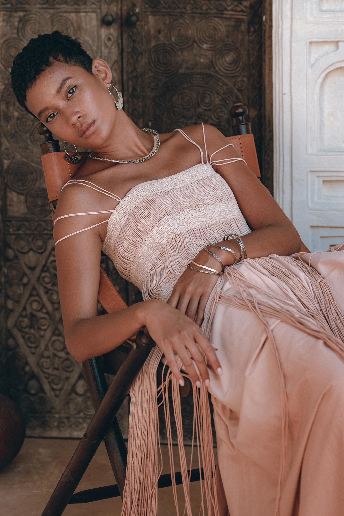 A woman with short hair sits in a wooden chair, wearing the AYA Sacred Wear Dusty Pink Tribal Goddess Slip Over net overlay dress.—an ethnic handwoven macrame layer up sheer net dress—posing gracefully by an ornate carved wooden door.