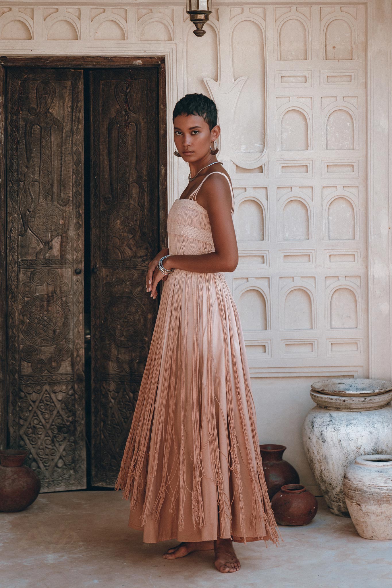 A woman with short hair stands barefoot by ornate wooden doors, wearing the AYA Sacred Wear Dusty Pink Tribal Goddess Slip Over net overlay dress. —an ethnic handwoven macrame, sheer net dress—amid pottery and white textured walls, creating a serene scene.