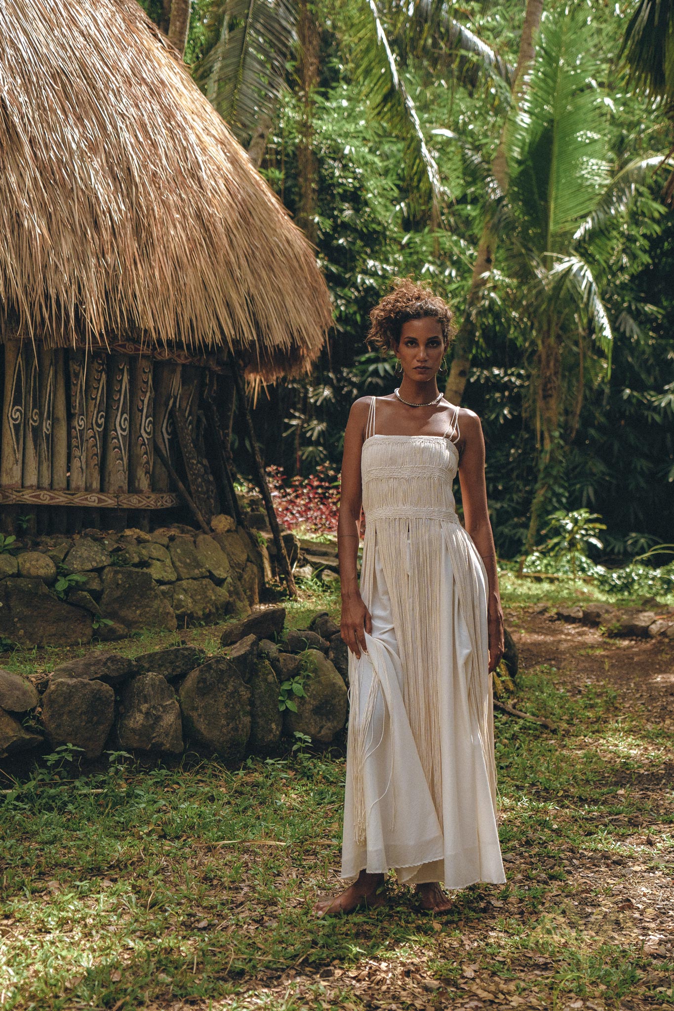 A woman in the AYA Sacred Wear Off-White Tribal Goddess Slip Over, an ethnic handwoven macrame sheer net dress, stands barefoot on grass by a rustic thatched hut amid lush tropical greenery.