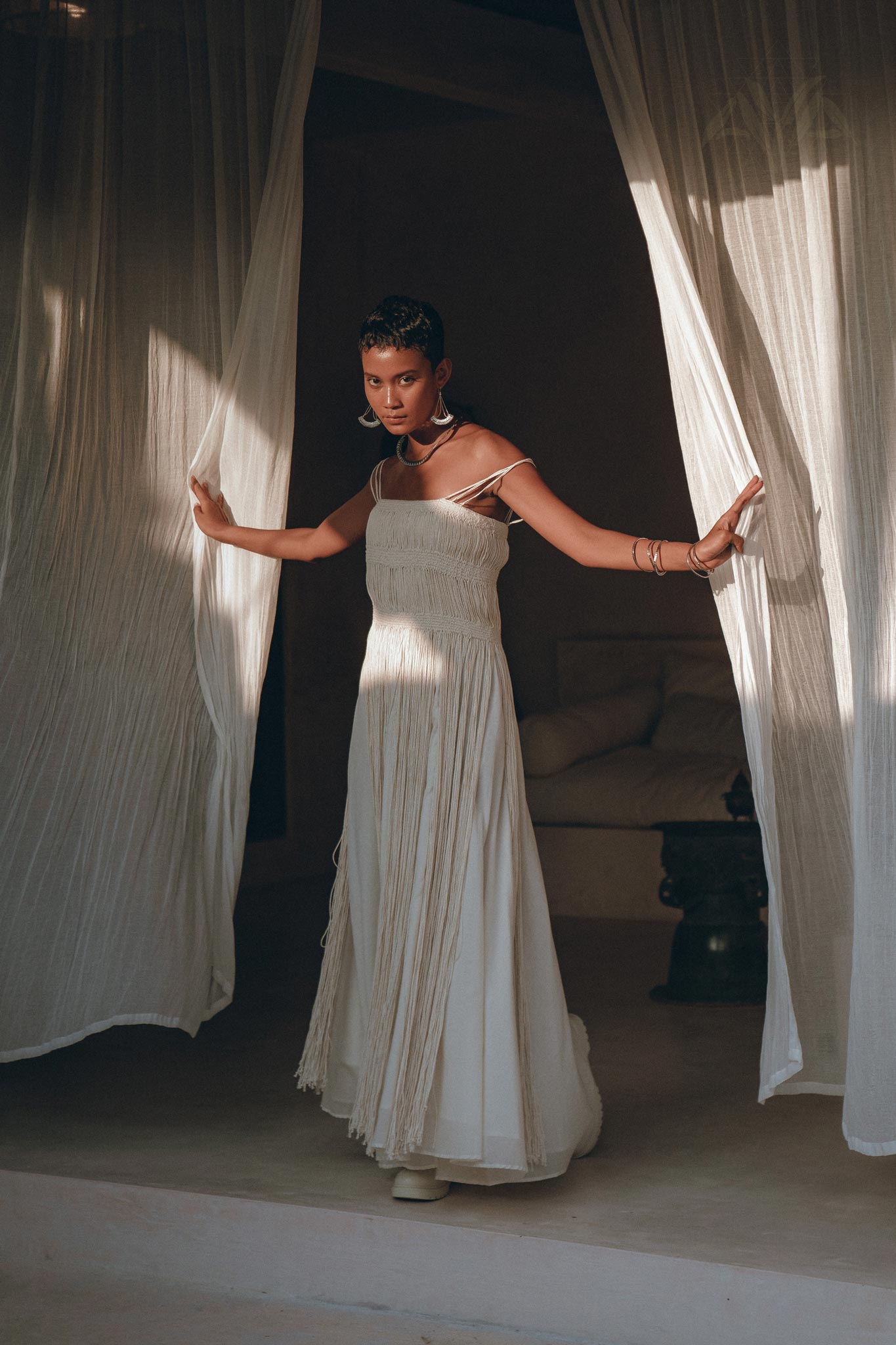 A woman in the Off-White Tribal Goddess Slip Over by AYA Sacred Wear stands in a doorway, holding sheer curtains as soft natural light illuminates her elegant pose and the serene, minimalist interior behind her.