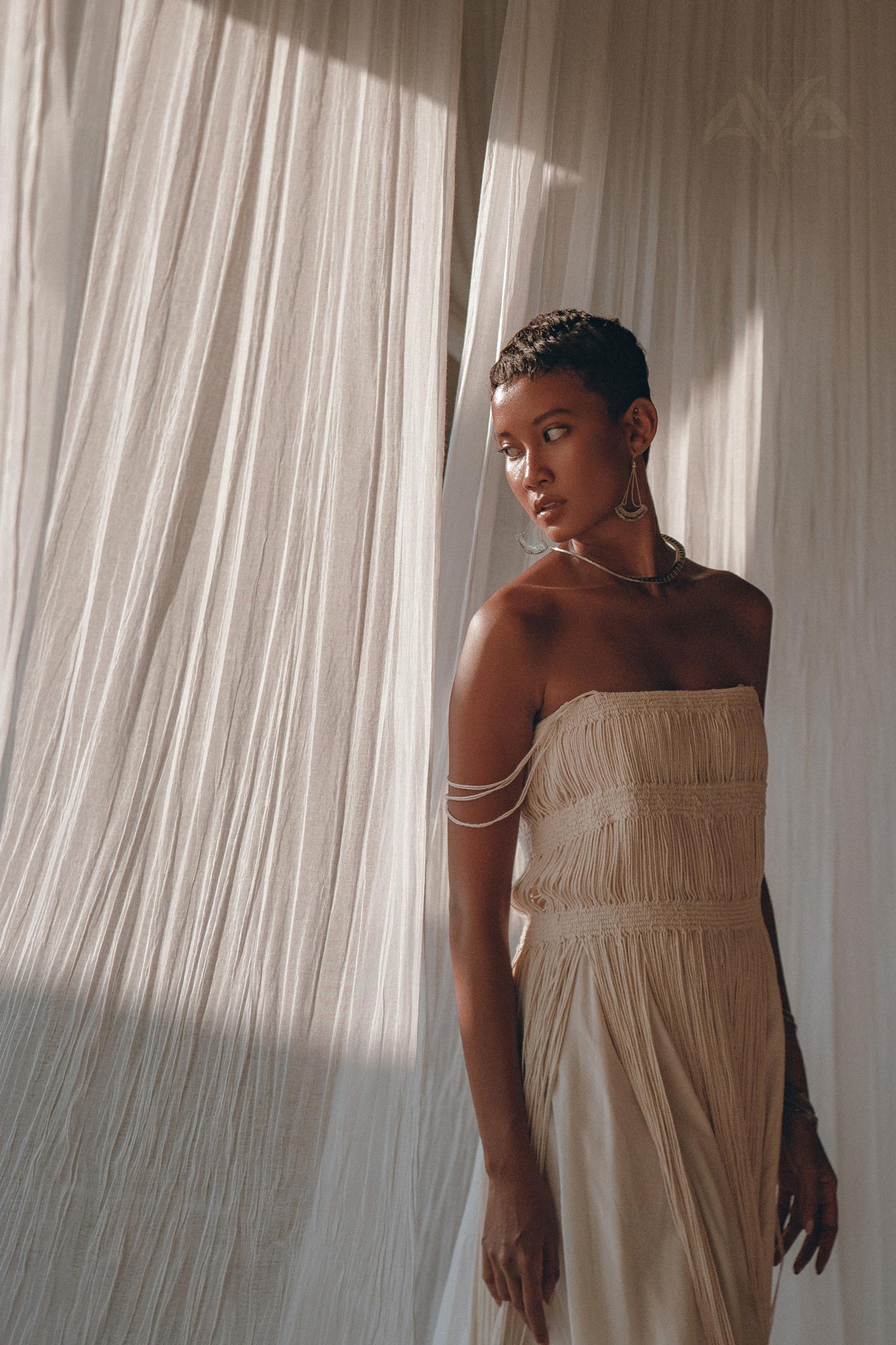 A woman in the AYA Sacred Wear Off-White Tribal Goddess Slip Over, an ethnic handwoven macrame sheer net layer-up, stands before cream curtains, gazing to the side as natural light highlights her look.