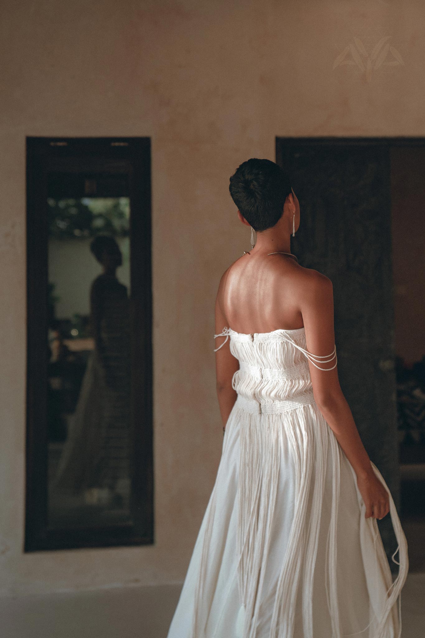 A woman wears the Off-White Tribal Goddess Slip Over by AYA Sacred Wear—an ethnic handwoven macrame layer-up sheer net dress. Indoors, she stands with her back to the camera, facing a dark doorway and softly lit reflective glass.