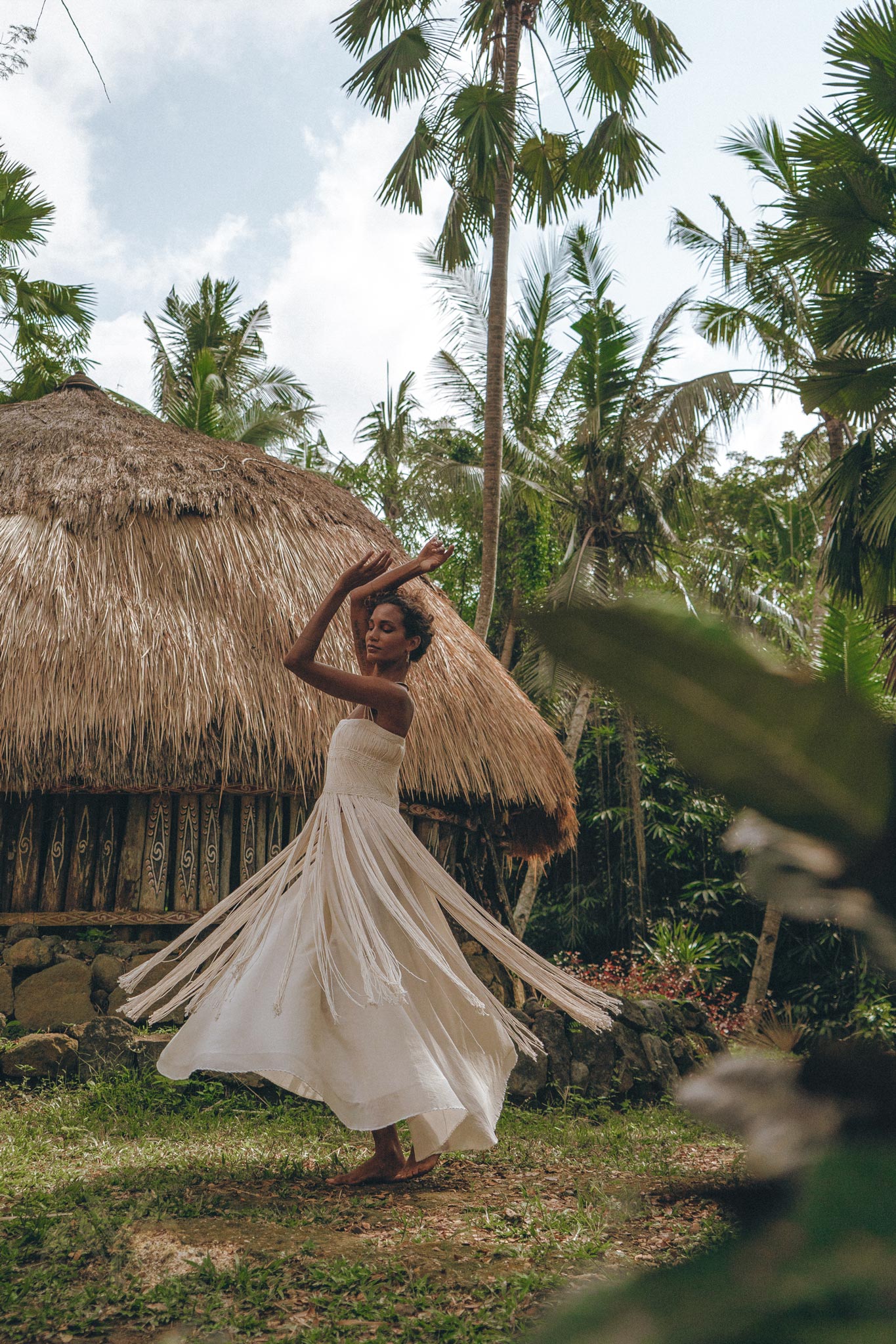 A woman in an Off-White Tribal Goddess Slip Over by AYA Sacred Wear dances joyfully outdoors near a thatched-roof hut, surrounded by lush palm trees and tropical greenery. The ethnic handwoven macrame layer adds a unique, sheer touch.