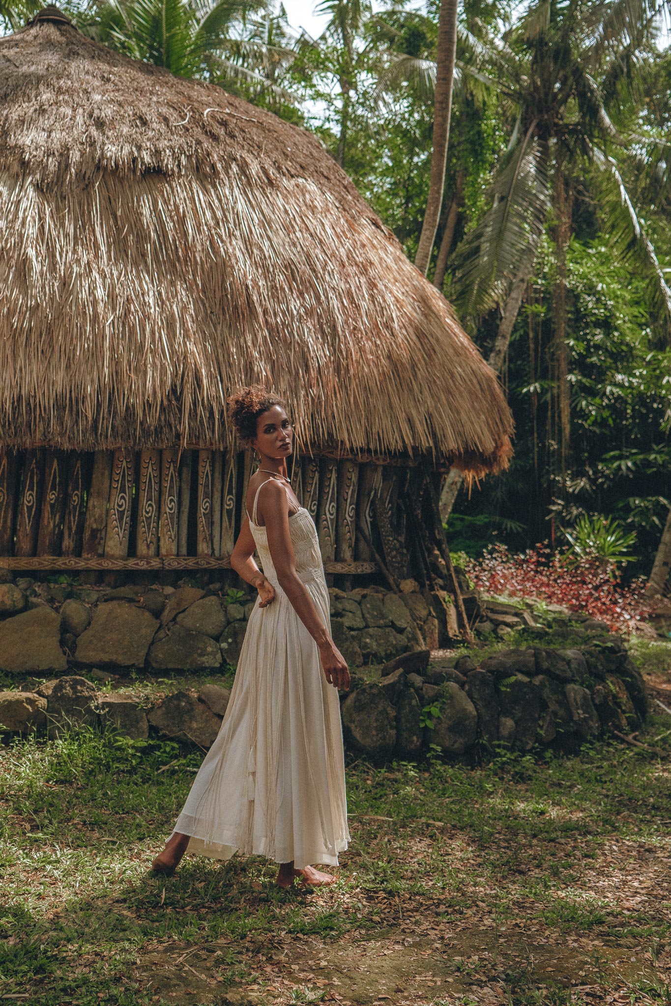 A woman wears the AYA Sacred Wear Off-White Tribal Goddess Slip Over, an ethnic handwoven macrame sheer net, standing before a thatched-roof hut amid lush greenery and palm trees.