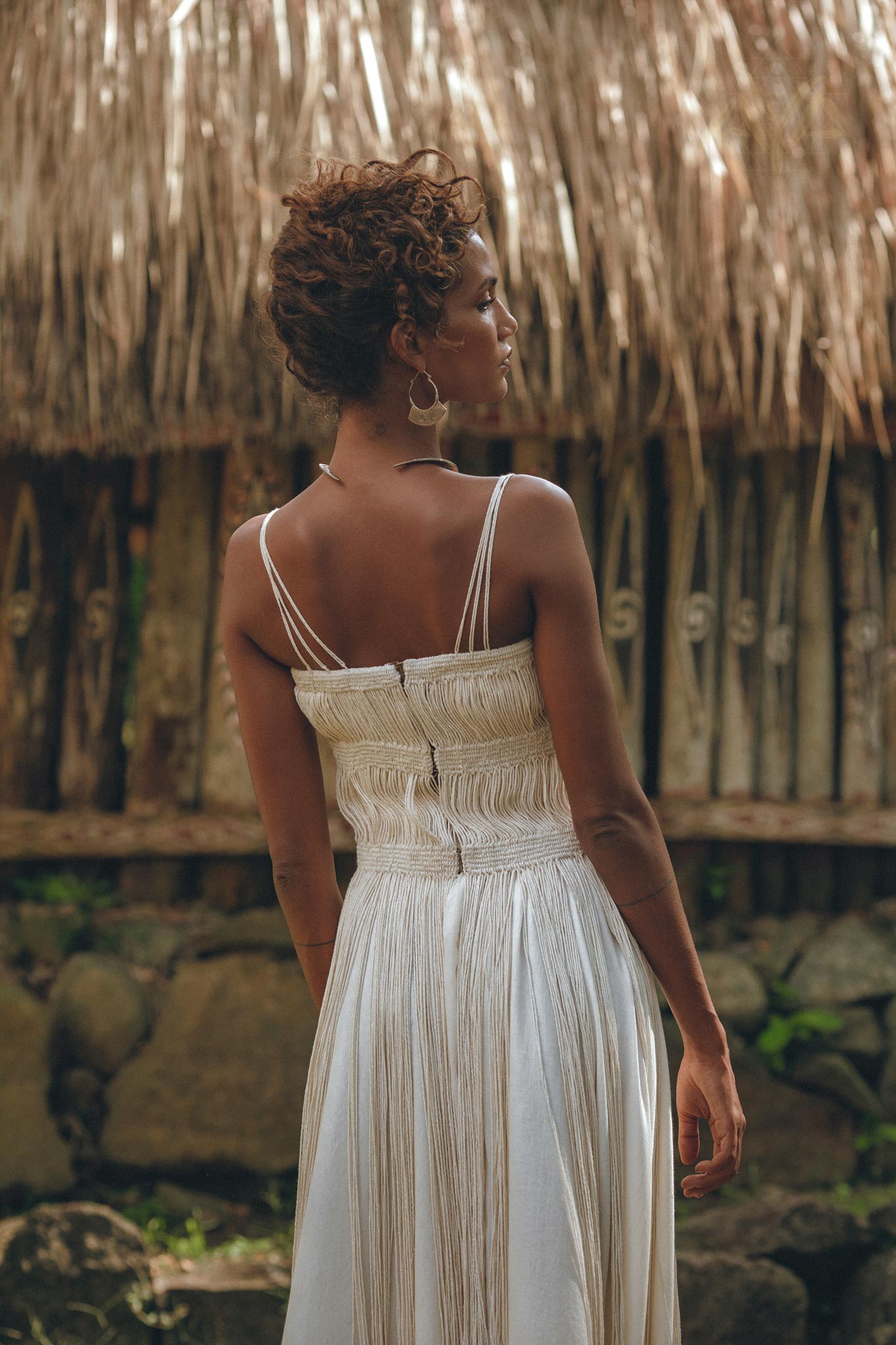 A woman in the Off-White Tribal Goddess Slip Over by AYA Sacred Wear stands outdoors, her back turned amid rustic wooden fencing, stones, and thatched roofing—a serene setting for this ethnic handwoven macrame layer up sheer net dress.
