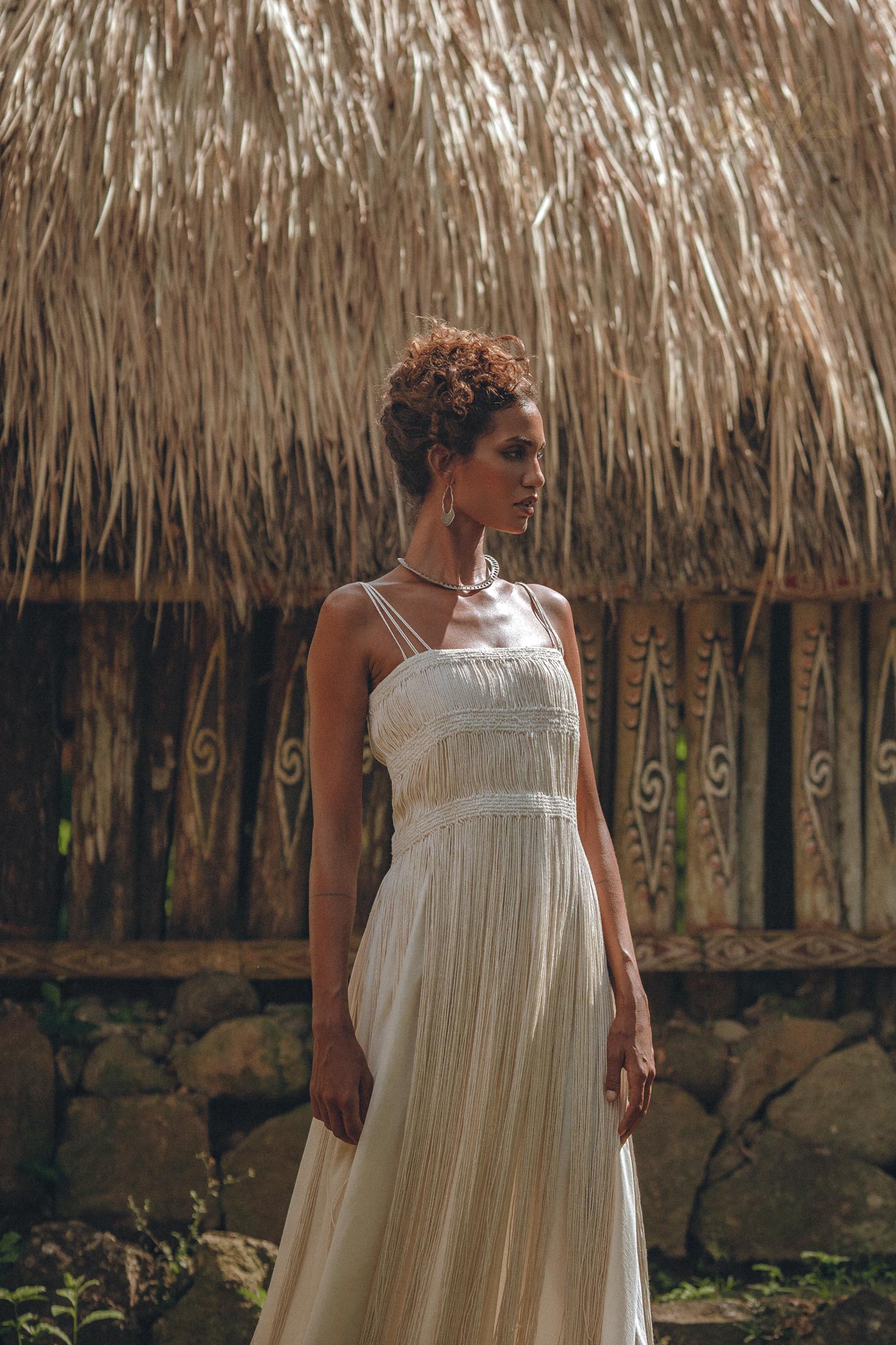 A woman wearing the Off-White Tribal Goddess Slip Over—an ethnic handwoven macrame layer up sheer net by AYA Sacred Wear—stands outdoors near a rustic thatched structure, sunlight highlighting her and the textured surroundings.