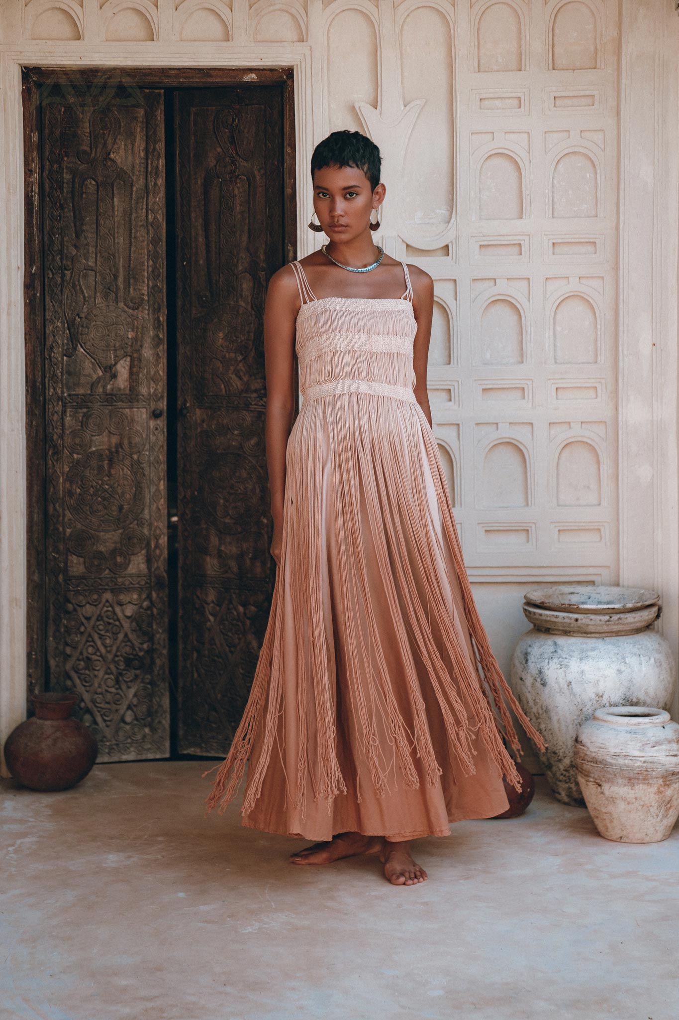 A woman in the AYA Sacred Wear Dusty Pink Tribal Goddess Slip Over—an ethnic handwoven macrame, sheer net dress—stands barefoot on a stone floor before carved wooden doors and cream-textured walls adorned with pottery.