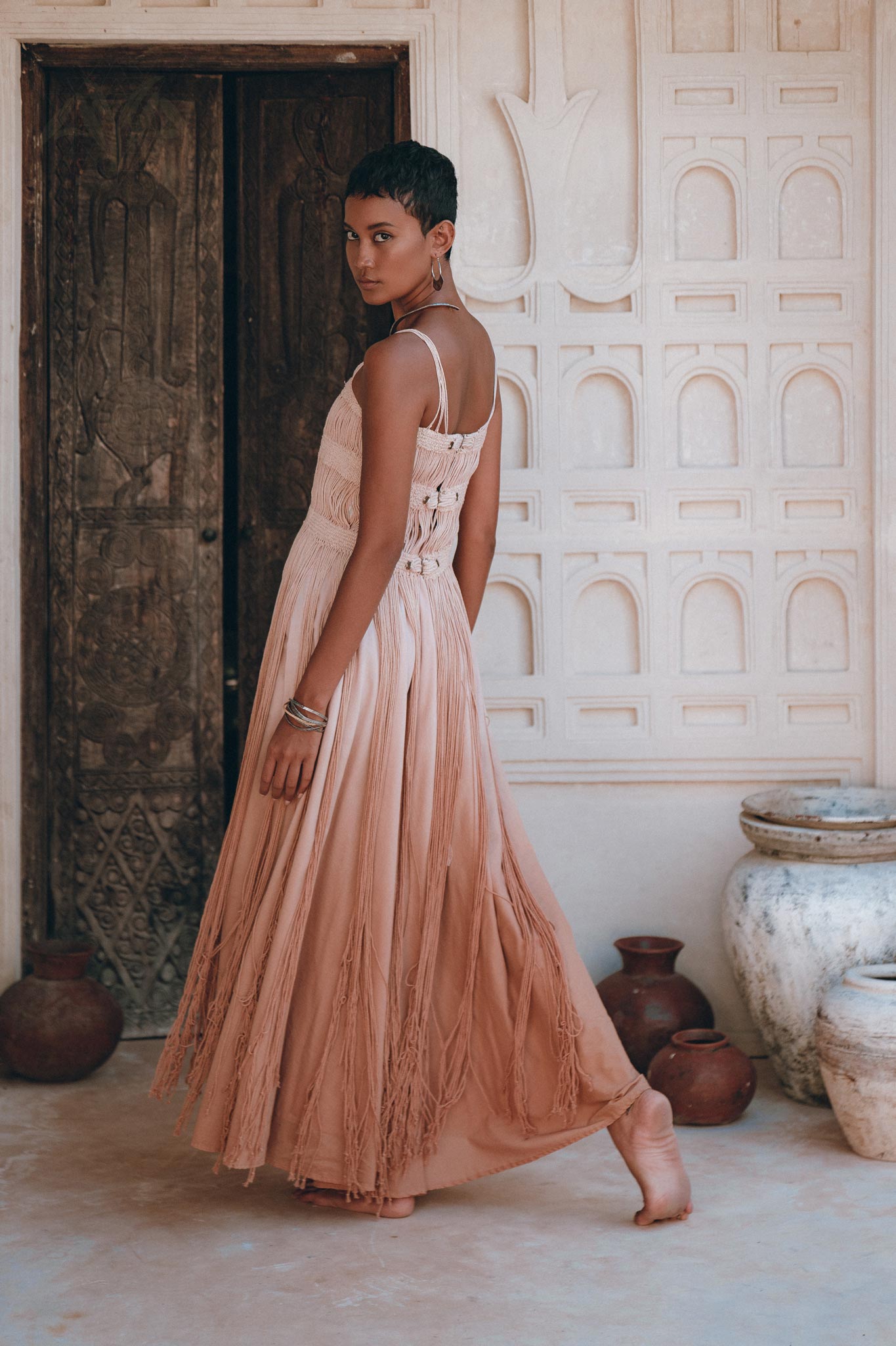 A woman with short hair, barefoot, stands indoors turning to look over her shoulder. She wears the AYA Sacred Wear Dusty Pink Tribal Goddess Slip Over—an ethnic handwoven macrame layer up sheer net—amid rustic pottery and carved walls.