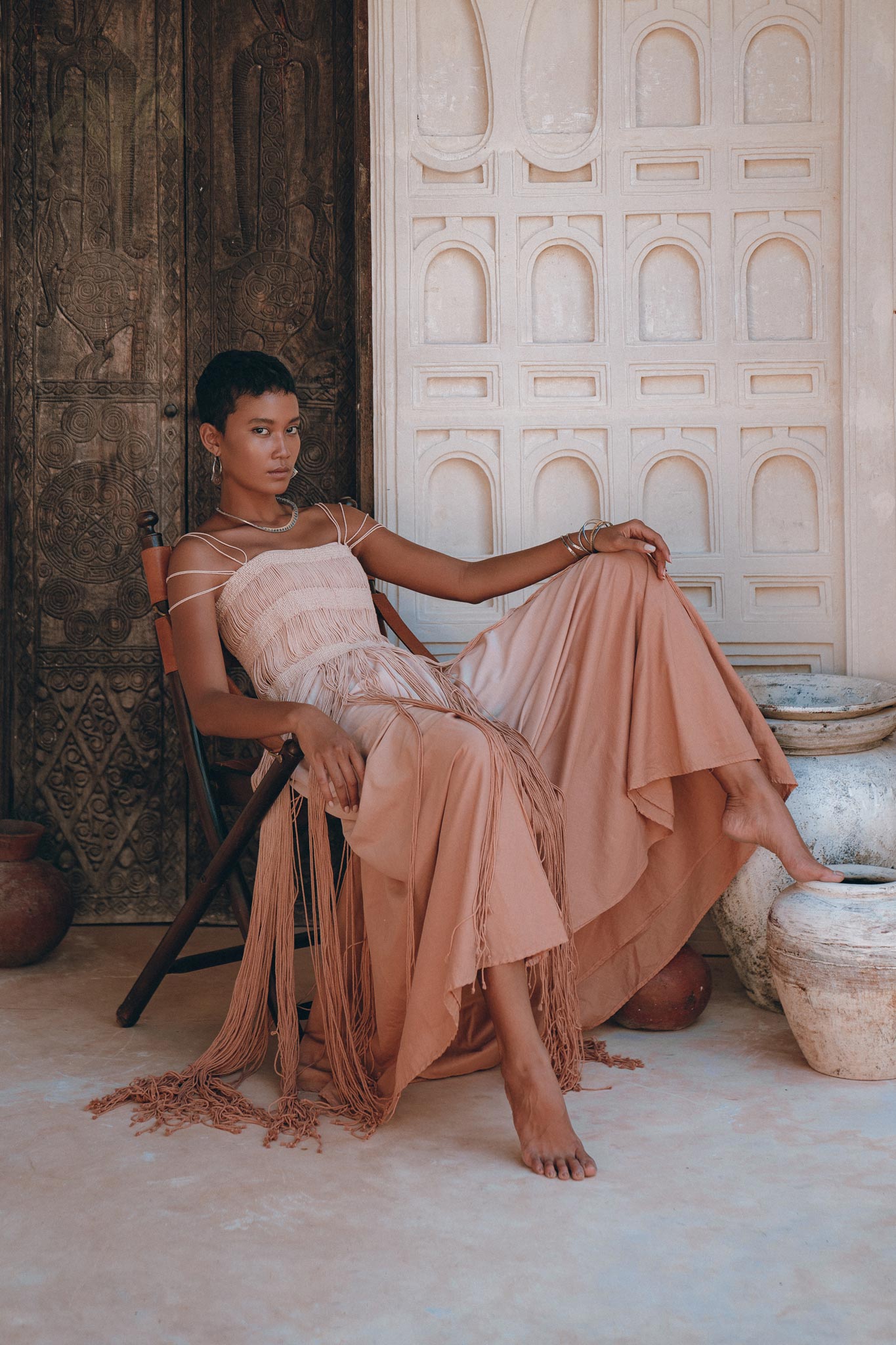 A person in a Dusty Pink Tribal Goddess Slip net overlay dress. by AYA Sacred Wear sits barefoot on a wooden chair, leaning back with one leg draped over the armrest, against an ornate light-colored wall and clay pottery.