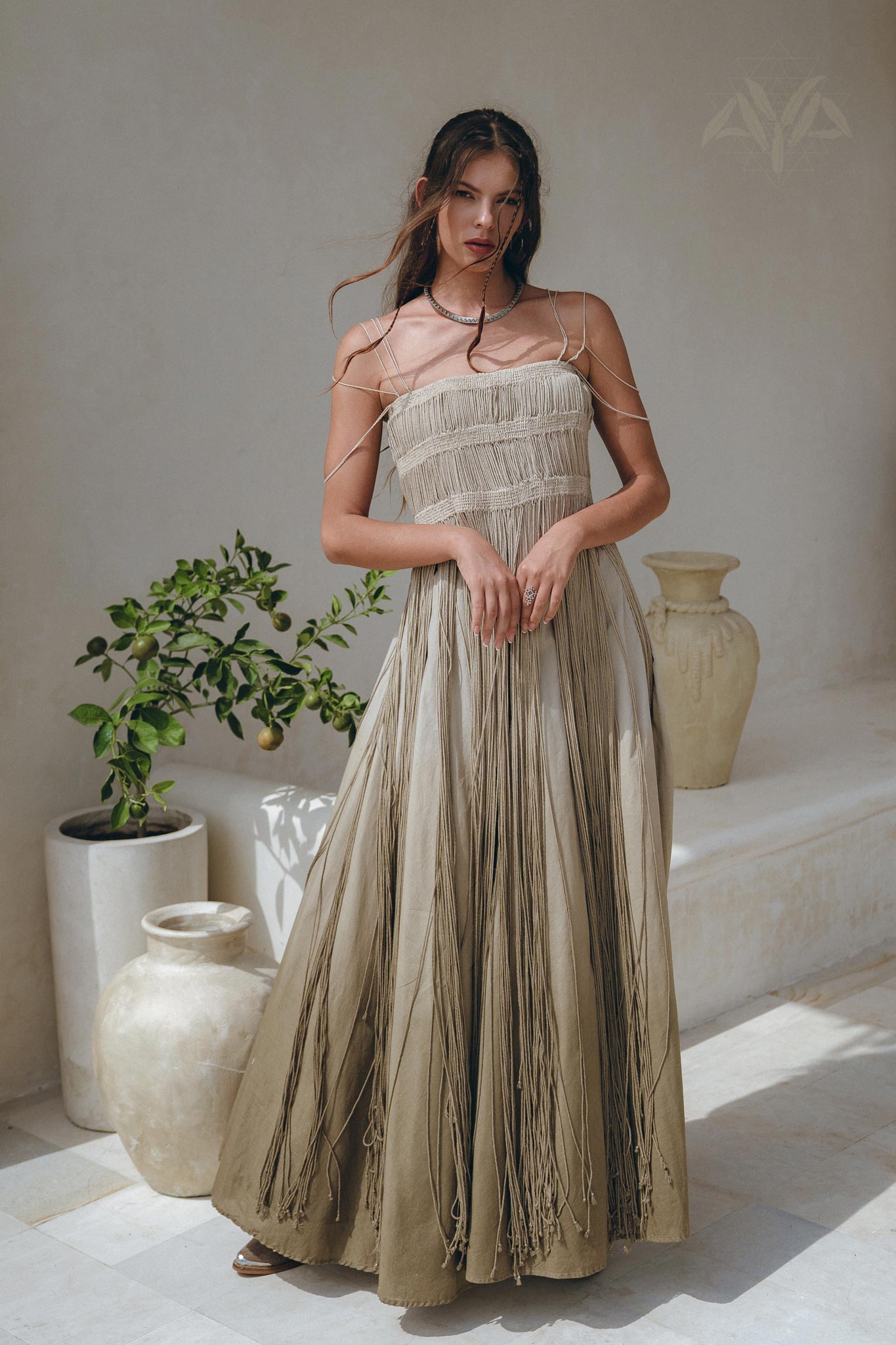 A woman stands indoors wearing the AYA Sacred Wear Sage Green Tribal Goddess Slip Over, an ethnic handwoven macrame layer-up sheer net dress, posing near pottery and a green plant against a light wall.