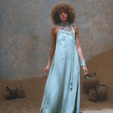 A woman stands indoors on sand, elegantly dressed in the Blue Aurora Greek Goddess Dress by AYA Sacred Wear. The flowing one-shoulder macrame toga maxi dress is complemented by her jewelry, while the background features large, weathered pots that enhance the tranquil and artistic ambiance.