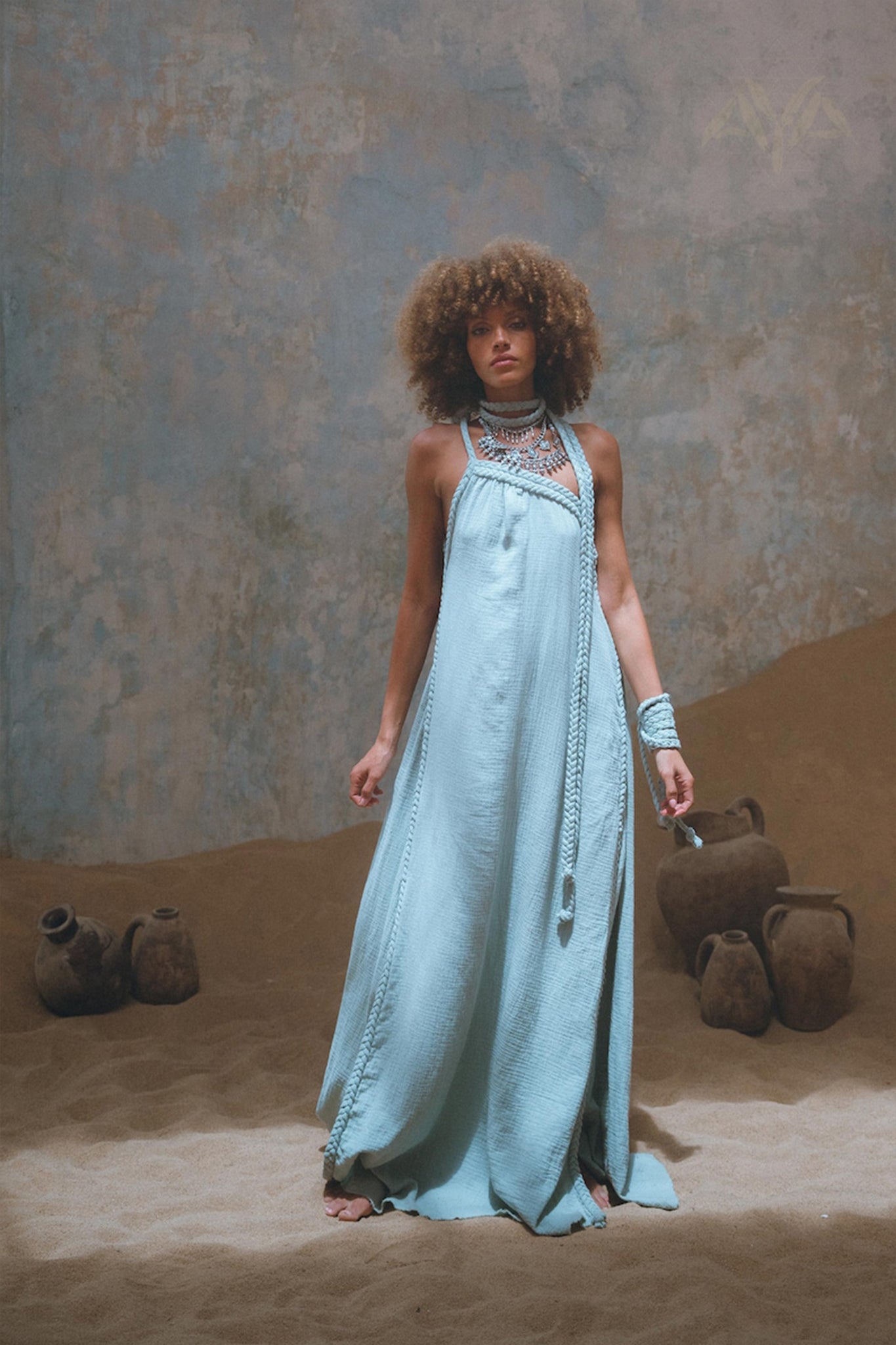 A woman stands indoors on sand, elegantly dressed in the Blue Aurora Greek Goddess Dress by AYA Sacred Wear. The flowing one-shoulder macrame toga maxi dress is complemented by her jewelry, while the background features large, weathered pots that enhance the tranquil and artistic ambiance.