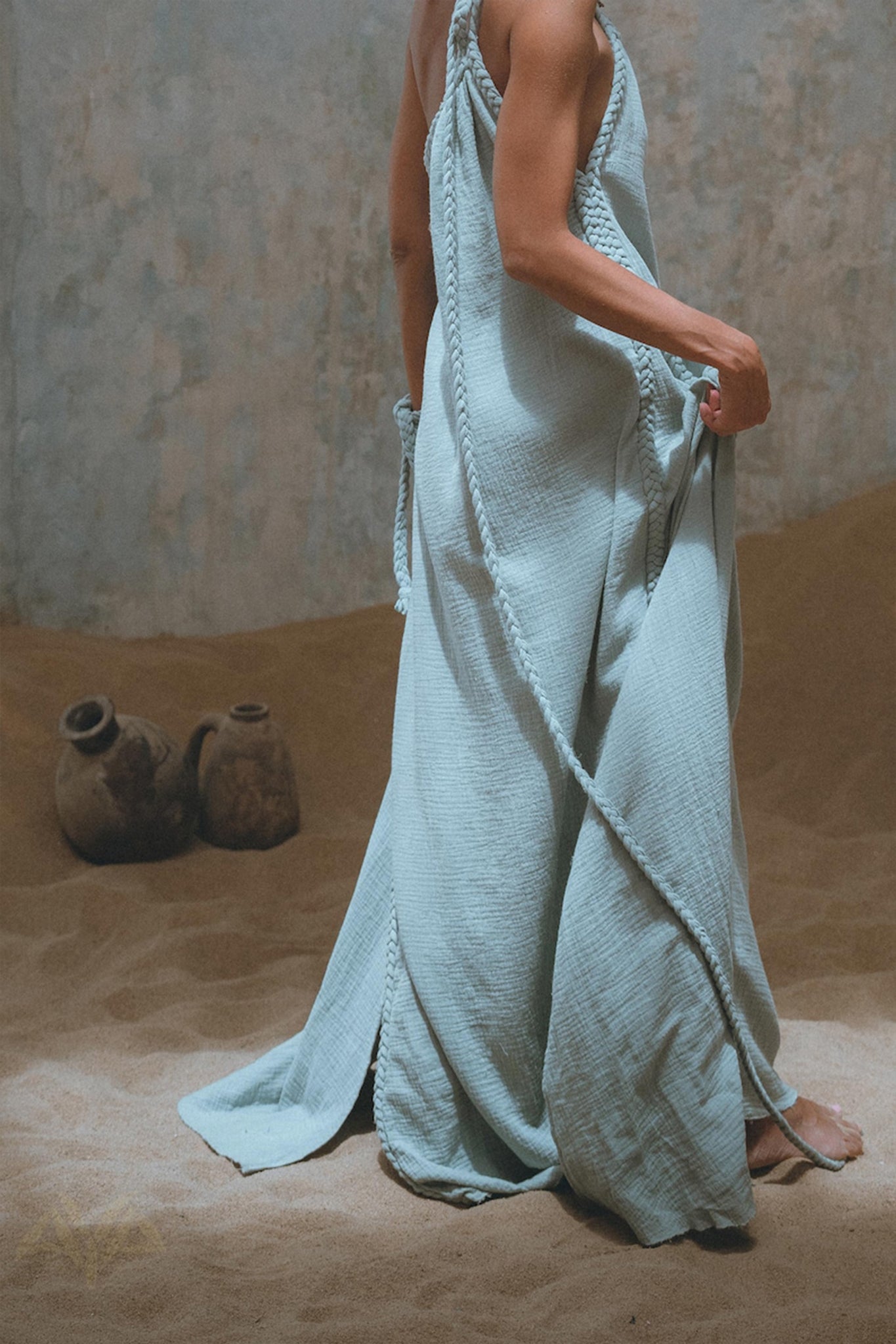 In the background of a sandy setting, a person elegantly wears AYA Sacred Wear's Blue Aurora Greek Goddess Dress. The flowing, light blue toga-style dress features intricate macrame and a one-shoulder design, complete with braided details, while two clay pots are visible on the ground.