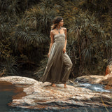 Barefoot on rocky ground by a stream, a woman in the Sage Green Tribal Goddess Slip Over from AYA Sacred Wear gazes thoughtfully, surrounded by lush foliage and trees. The ethnic handwoven macrame layer shines in the natural setting.