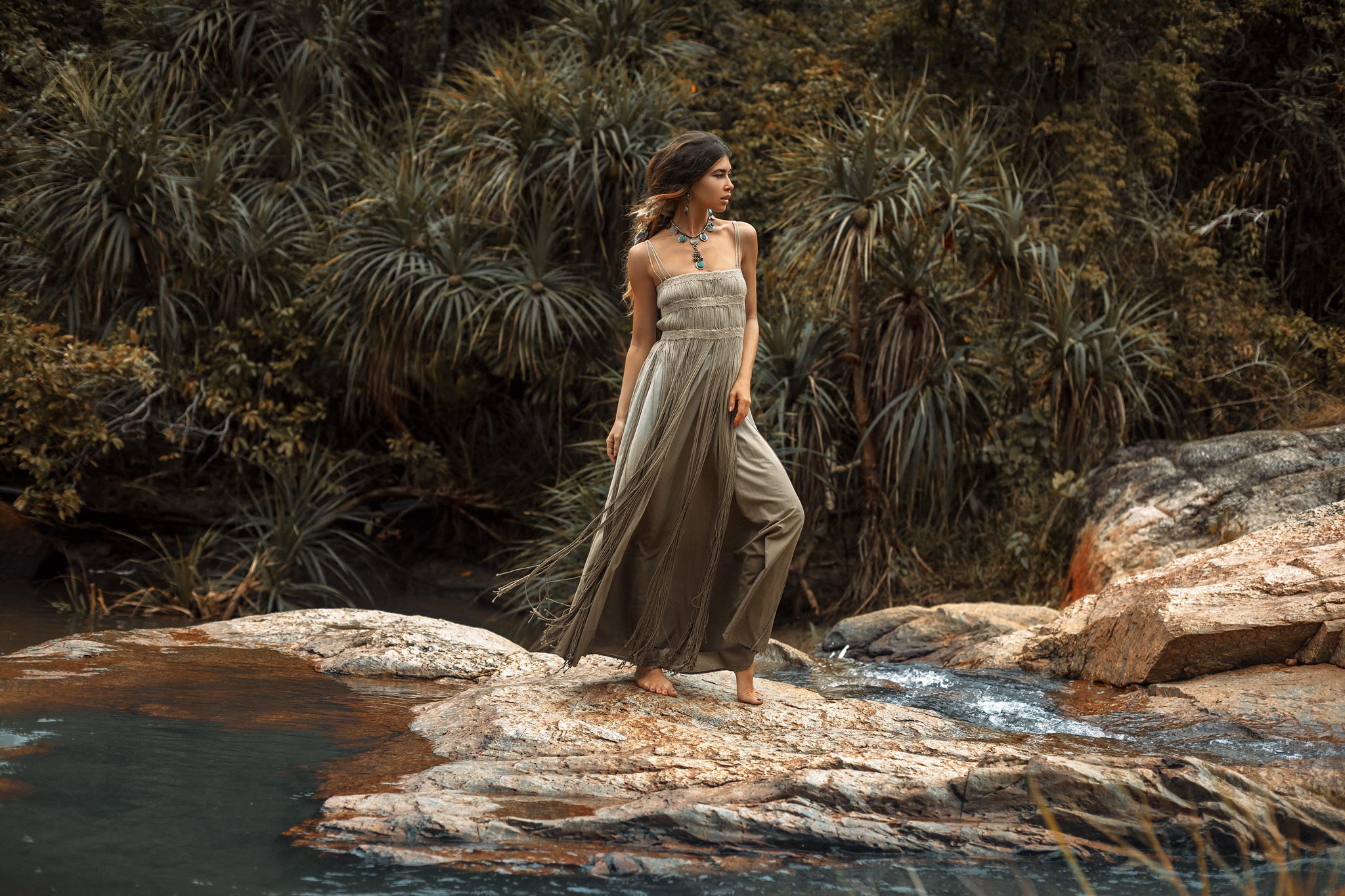 Barefoot on rocky ground by a stream, a woman in the Sage Green Tribal Goddess Slip Over from AYA Sacred Wear gazes thoughtfully, surrounded by lush foliage and trees. The ethnic handwoven macrame layer shines in the natural setting.