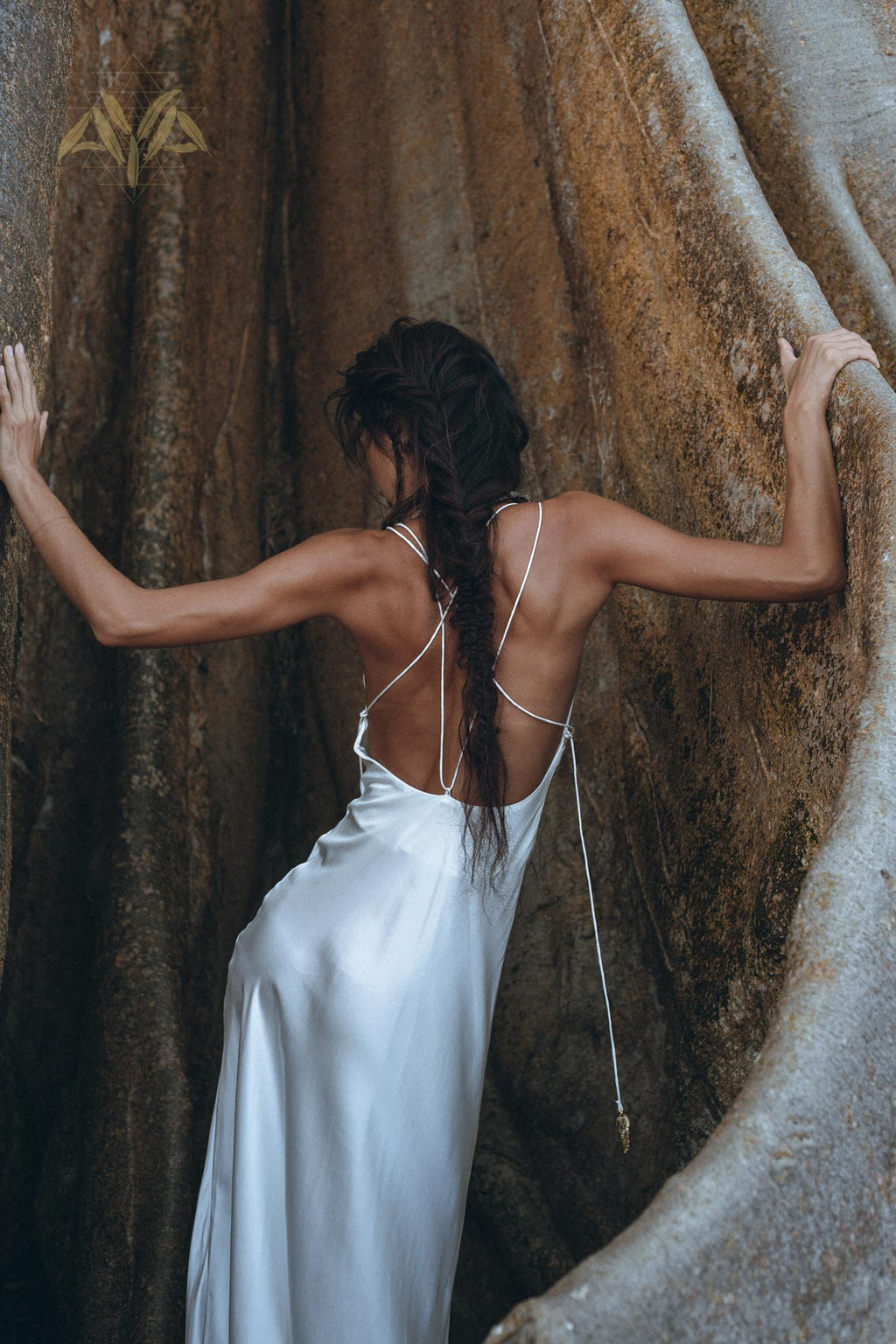 A woman wearing the White Peace Silk Simple Wedding Dress by AYA Sacred Wear stands between two large, smooth rocks. Her long hair is styled in a braid that falls down her back as she faces away, touching the rocks with both hands. The scene is serene and natural.