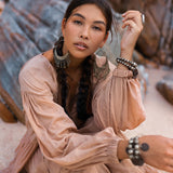 A woman with braided hair sits on rocky terrain, wearing a flowing Dusty Pink Bohemian Prom Dress from AYA Sacred Wear. She accessorizes with large earrings, bracelets, and rings. Her calm and serene expression complements the natural, earthy background.