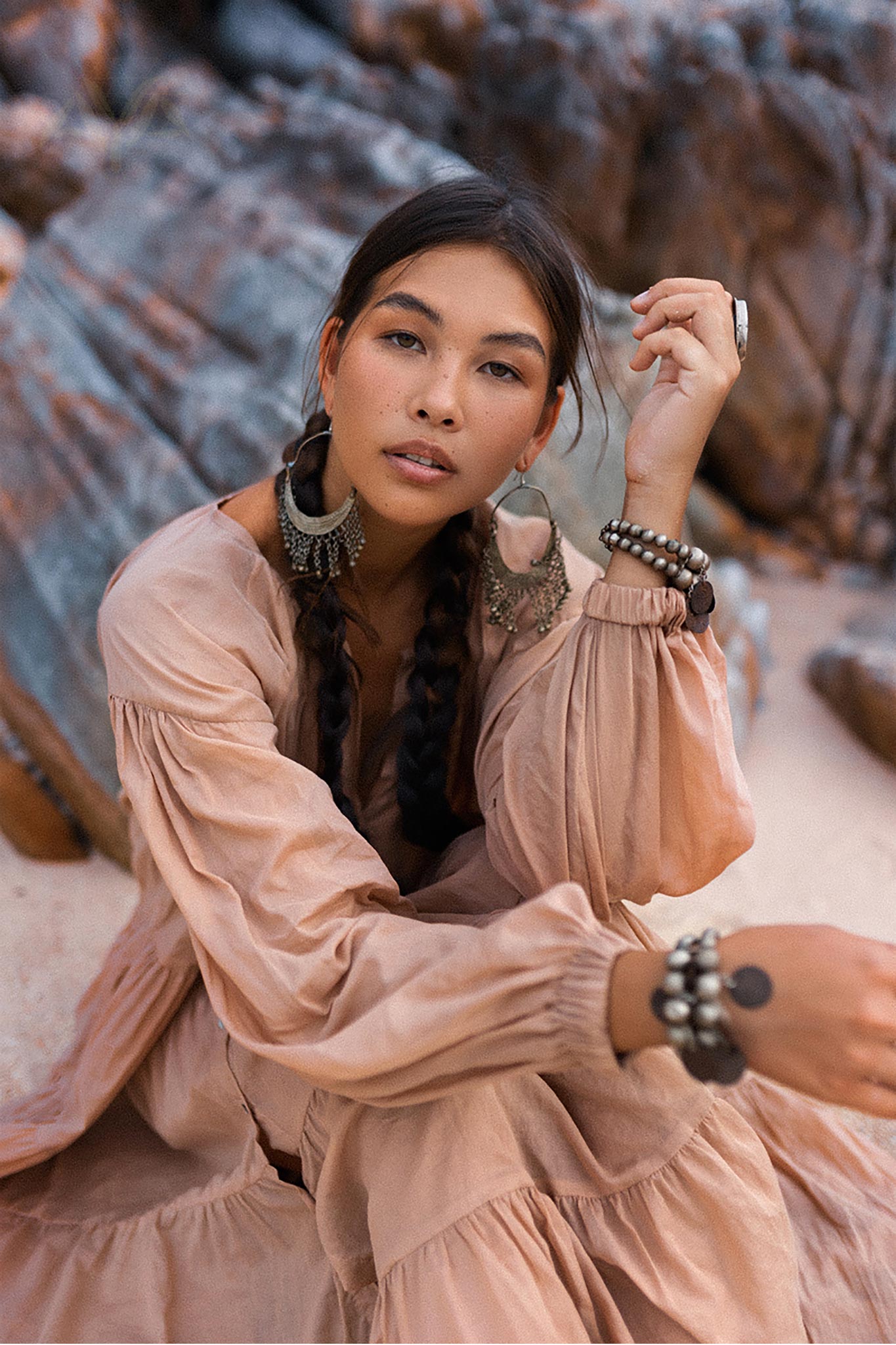 A woman with braided hair sits on rocky terrain, wearing a flowing Dusty Pink Bohemian Prom Dress from AYA Sacred Wear. She accessorizes with large earrings, bracelets, and rings. Her calm and serene expression complements the natural, earthy background.