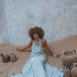 A person with curly hair sits on the sand against a textured wall, wearing the Blue Aurora Greek Goddess Dress from AYA Sacred Wear, a flowing macrame toga dress featuring an elegant one-shoulder design. Layered necklaces adorn them, while rustic clay pots are scattered around, enhancing the earthy and serene atmosphere.