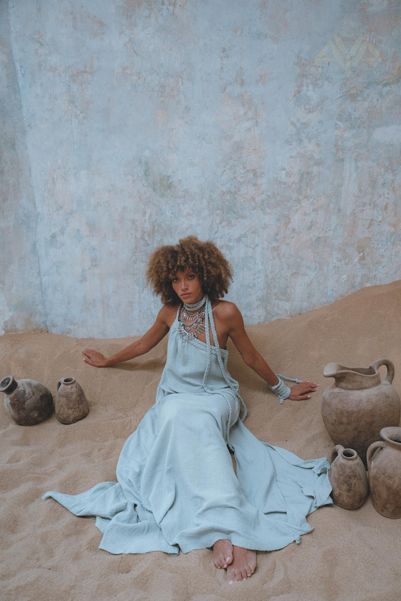 A person with curly hair sits on the sand against a textured wall, wearing the Blue Aurora Greek Goddess Dress from AYA Sacred Wear, a flowing macrame toga dress featuring an elegant one-shoulder design. Layered necklaces adorn them, while rustic clay pots are scattered around, enhancing the earthy and serene atmosphere.