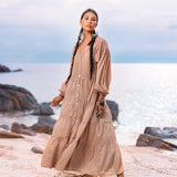 A woman with long braided hair stands on a sandy beach, wearing AYA Sacred Wear's Boho Dress for Women, a flowing dusty pink bohemian prom dress. Crafted from soft cotton fabric, it complements the serene ocean and rocks behind her, with a gentle, cloudy sky above.