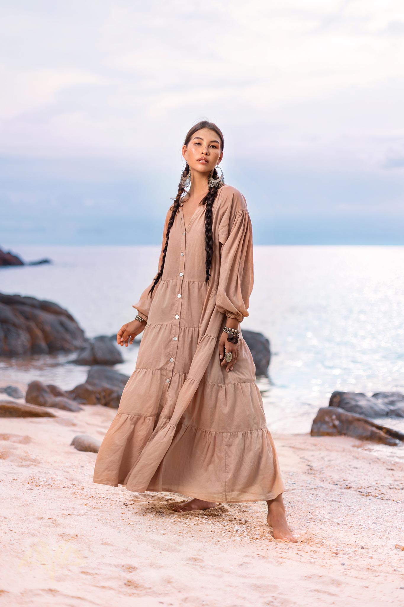 A woman with long braided hair stands on a sandy beach, wearing AYA Sacred Wear's Boho Dress for Women, a flowing dusty pink bohemian prom dress. Crafted from soft cotton fabric, it complements the serene ocean and rocks behind her, with a gentle, cloudy sky above.