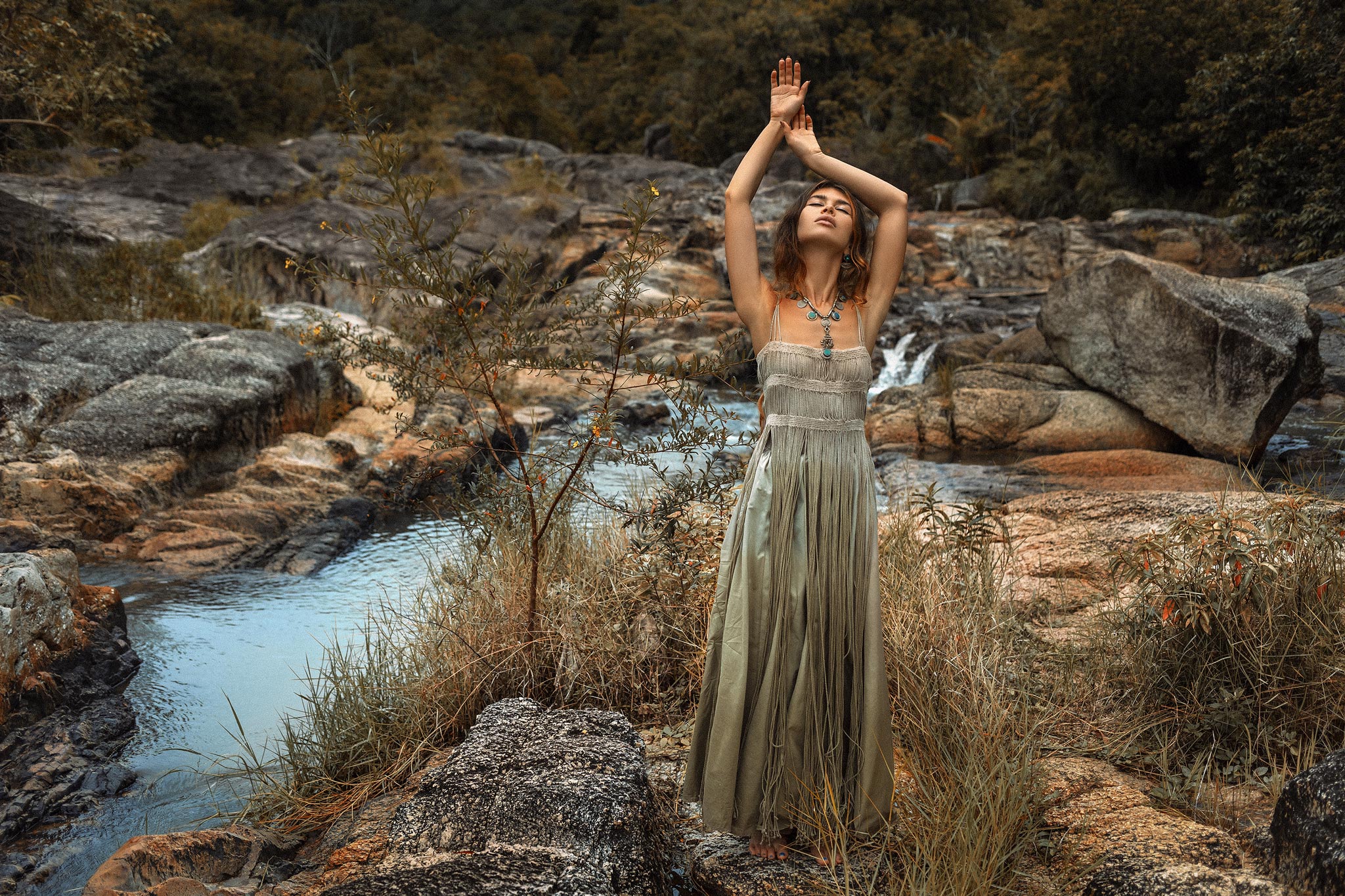 A woman in the Sage Green Tribal Goddess Slip Over by AYA Sacred Wear stands on rocks beside a stream, surrounded by lush greenery and boulders, her arms gracefully raised overhead.