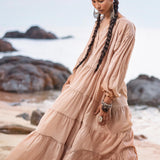 A woman with long braided hair wears an AYA Sacred Wear Boho Dress for Women in dusty pink. She stands thoughtfully on a rocky beach. Large earrings and bracelets complement her look, with the sea and a cloudy sky visible in the distance.