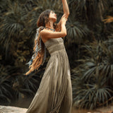 A barefoot woman poses gracefully on rocks in the AYA Sacred Wear Sage Green Tribal Goddess Slip Over—an ethnic handwoven macrame layer-up with sheer net—surrounded by lush green foliage for a serene, natural feel.