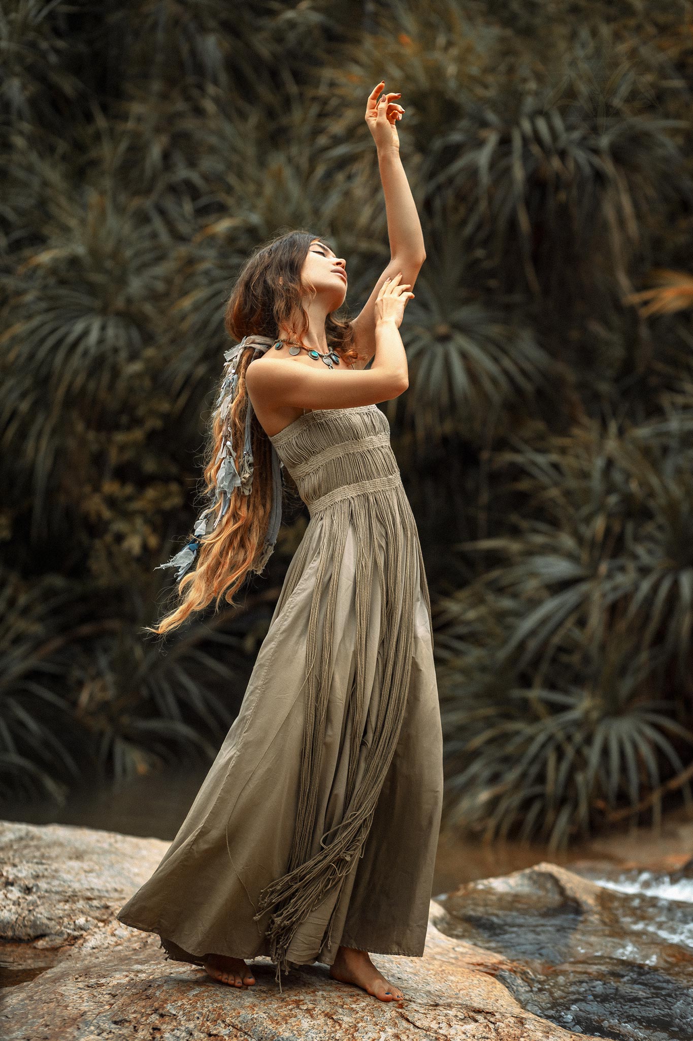 A barefoot woman poses gracefully on rocks in the AYA Sacred Wear Sage Green Tribal Goddess Slip Over—an ethnic handwoven macrame layer-up with sheer net—surrounded by lush green foliage for a serene, natural feel.