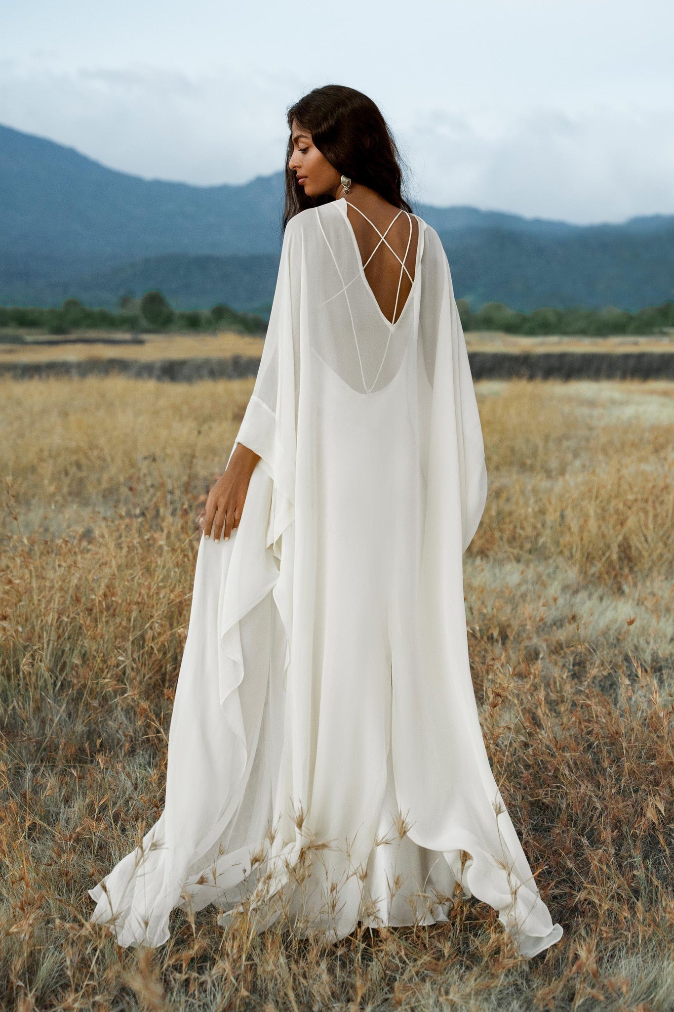 A woman with long dark hair wears the Mahadevi Dress in cream by AYA Sacred Wear, a pure silk chiffon goddess gown with crisscross back straps, standing in a dry grassy field with mountains and a cloudy sky behind her.