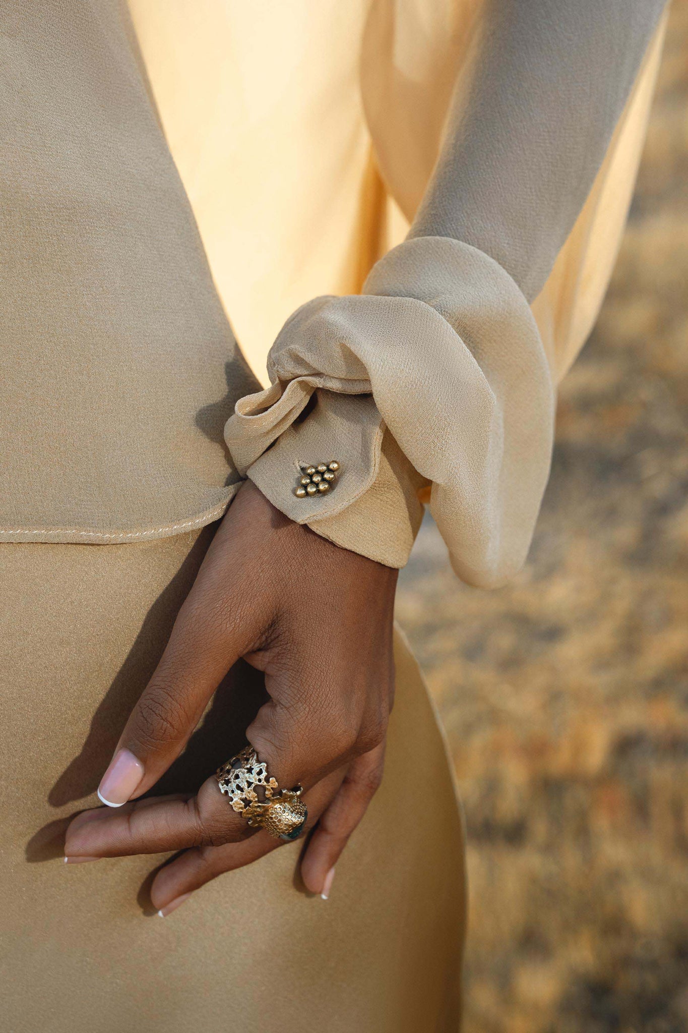 A close-up of a hand rests on the AYA Sacred Wear Maharani Blouse in Inka Gold—a pure silk chiffon top with sheer, flowy fabric and gold buttons. A statement gold ring shines, while the softly blurred background adds elegance.
