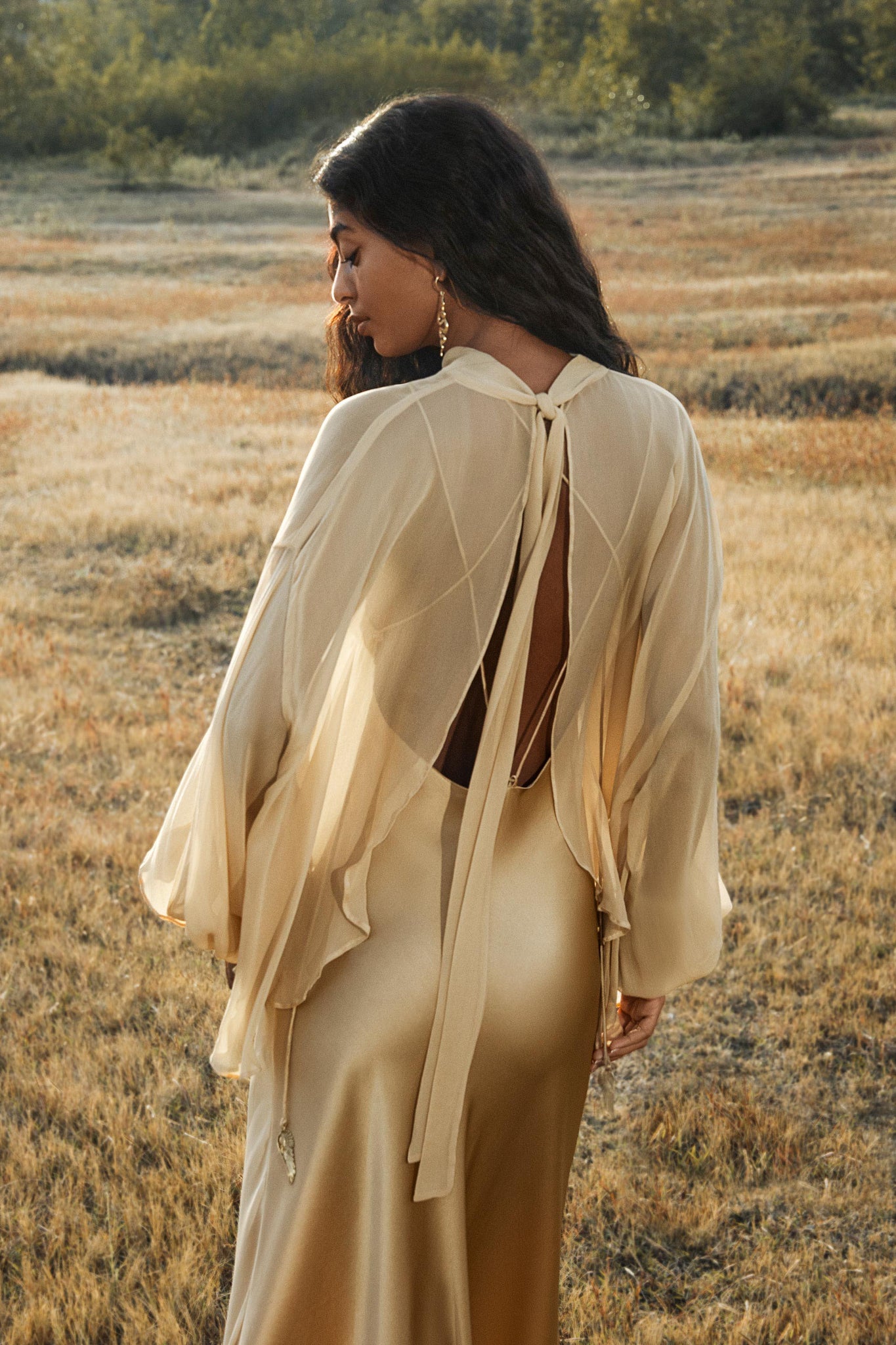 A woman with long hair stands in a dry, grassy field wearing the AYA Sacred Wear Maharani Blouse in Inka Gold—an elegant pure silk chiffon top. She gazes over her shoulder in the serene, sunlit setting.