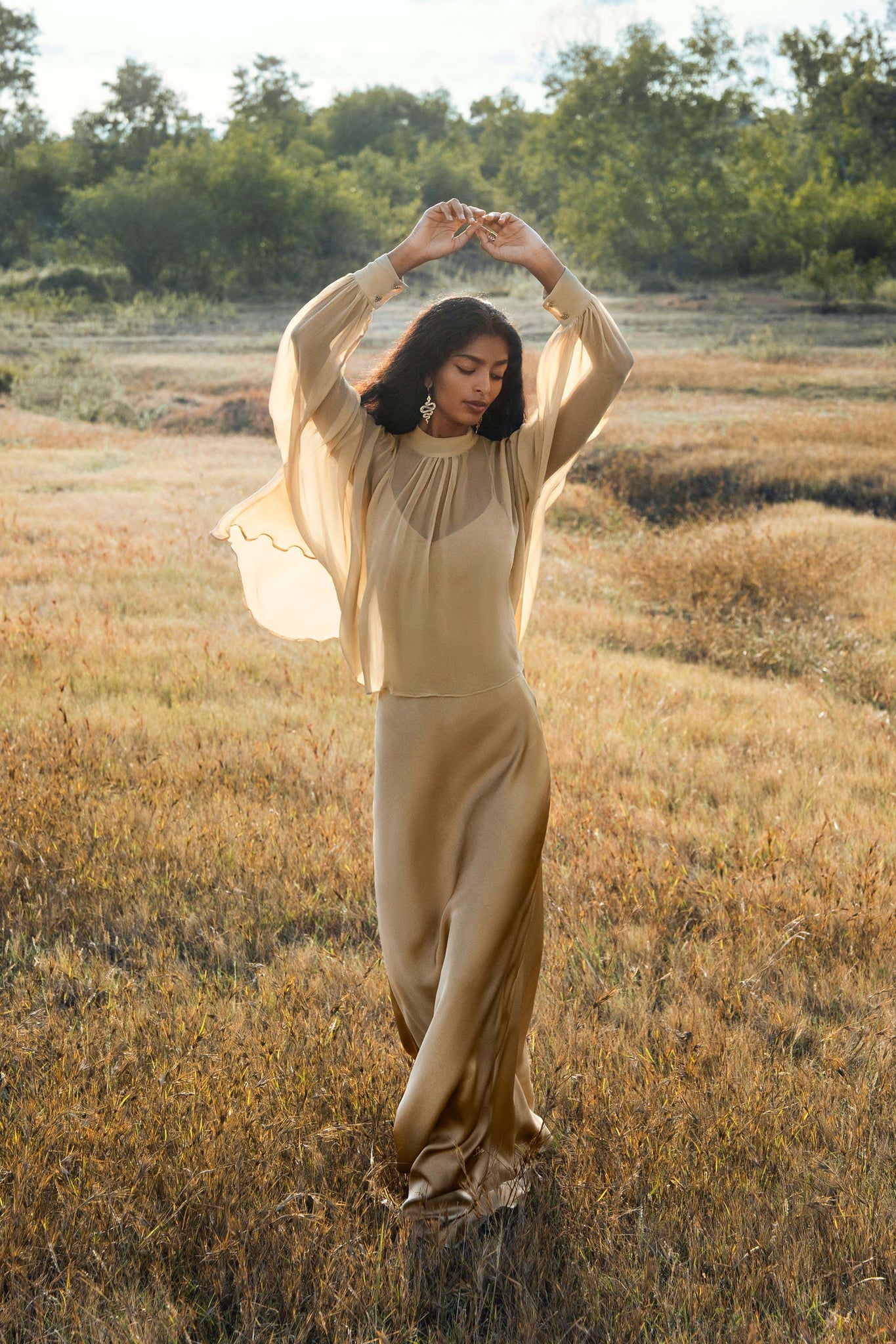 A woman in the AYA Sacred Wear Maharani Blouse Inka Gold Colour—an elegant pure silk chiffon top—stands in a sunlit field, arms raised and eyes closed, surrounded by tall grass and lush greenery.