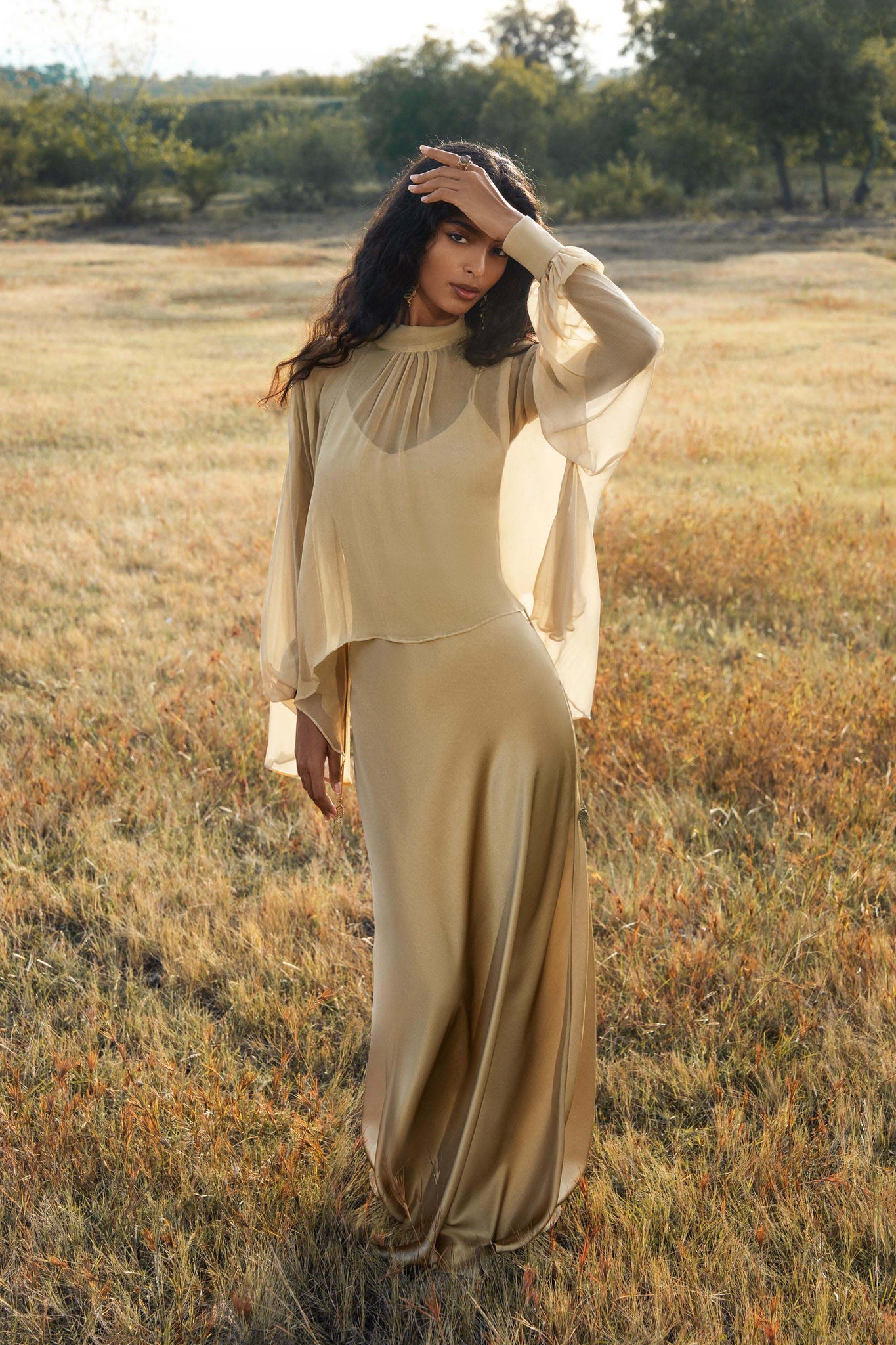 A woman stands in a sunlit field wearing the AYA Sacred Wear Maharani Blouse in Inka Gold—an elegant pure silk chiffon top with sheer sleeves—her long, wavy hair glowing as she gazes softly at the camera, trees framing the background.